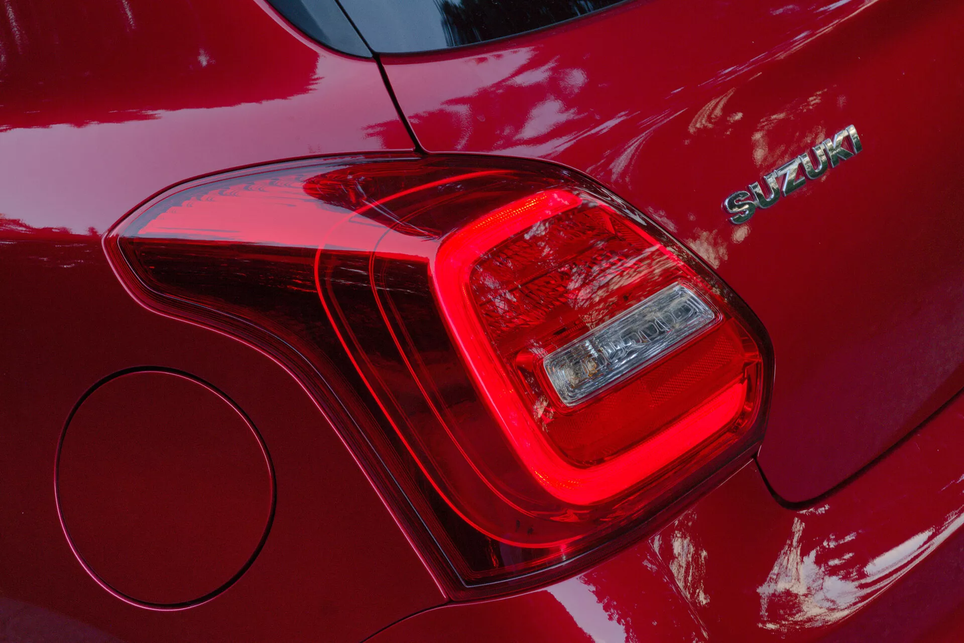 Primer plano del conjunto de luces traseras del lado izquierdo de un Suzuki Swift Sport 2017, resaltando el diseño segmentado de las unidades de luz, incluyendo la luz de freno y la luz de giro o reversa. La carrocería de color rojo profundo y acabado brillante refleja la luz y el entorno. Junto a la luz trasera se ve un emblema cromado con el nombre 'SWIFT'. La imagen también revela una tapa de combustible circular y muestra un lenguaje de diseño moderno y elegante con líneas aerodinámicas y materiales de construcción duraderos.