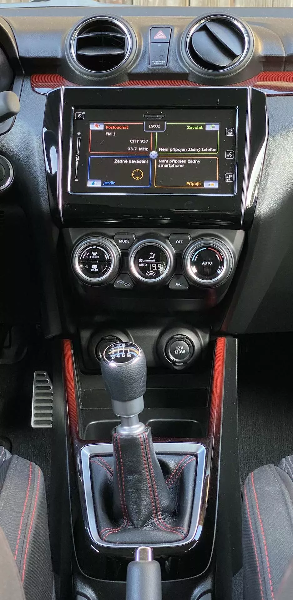 La consola central del interior de un Suzuki Swift Sport 2017, con un sistema de infoentretenimiento con botones físicos, ventilas circulares, un panel de control del clima manual con tres diales para temperatura, velocidad del ventilador y distribución del aire, y una palanca de cambios manual deportiva. La consola incluye una funda de cuero, un adorno decorativo con un posible patrón de fibra de carbono y una línea de acento rojo deportivo. Controles adicionales debajo de la palanca de cambios sugieren ayudas de conducción avanzadas.