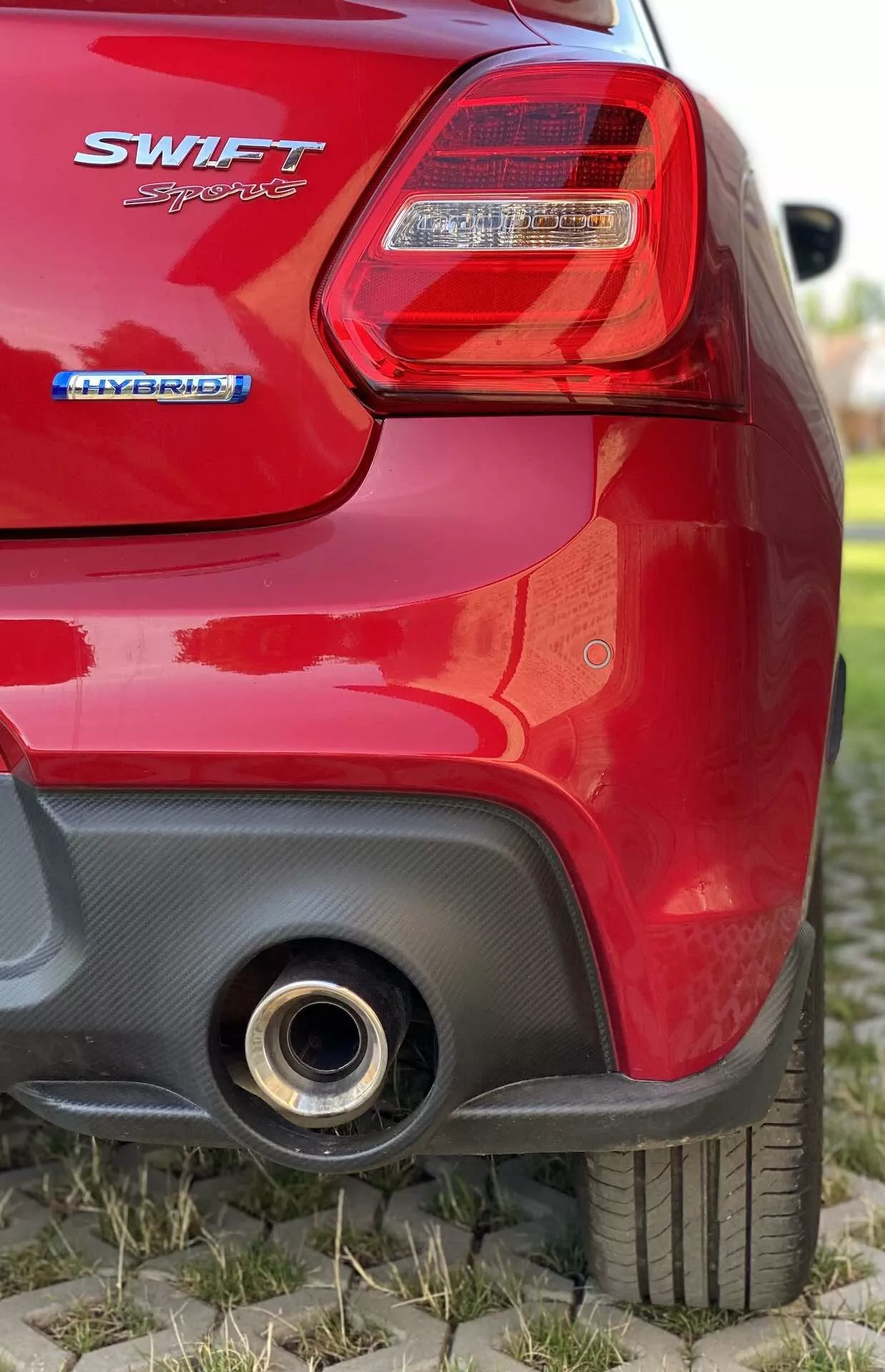 Vista de cerca de la sección trasera de un hatchback rojo Suzuki Swift 2017 desde un ángulo bajo. Imagen se enfoca en la luz trasera derecha con lente transparente y secciones de iluminación roja, insignia 'SWIFT' debajo de la luz, insignia 'HYBRID' debajo indicando tren motriz híbrido, parte trasera parcialmente desenfocada, neumático trasero derecho con patrón de la banda de rodadura visible, sección trasera en contraste negro con posible toma aerodinámica y tubo de escape metálico redondo en la parte inferior derecha que refleja luz.
