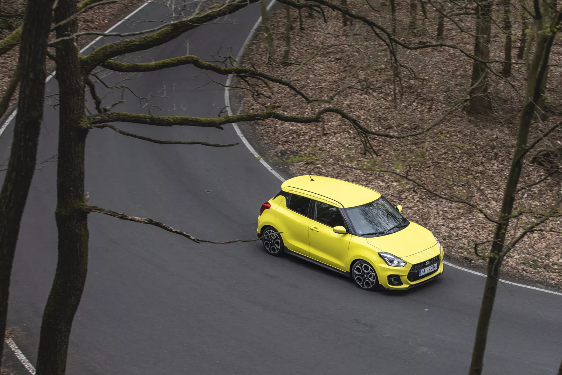 Vista aérea de un Suzuki Swift 1995 amarillo brillante con esquema de color de dos tonos viajando por una carretera serpenteante a través de un bosque, mostrando ruedas de aleación, faros delanteros LED modernos y un diseño compacto tipo hatchback.