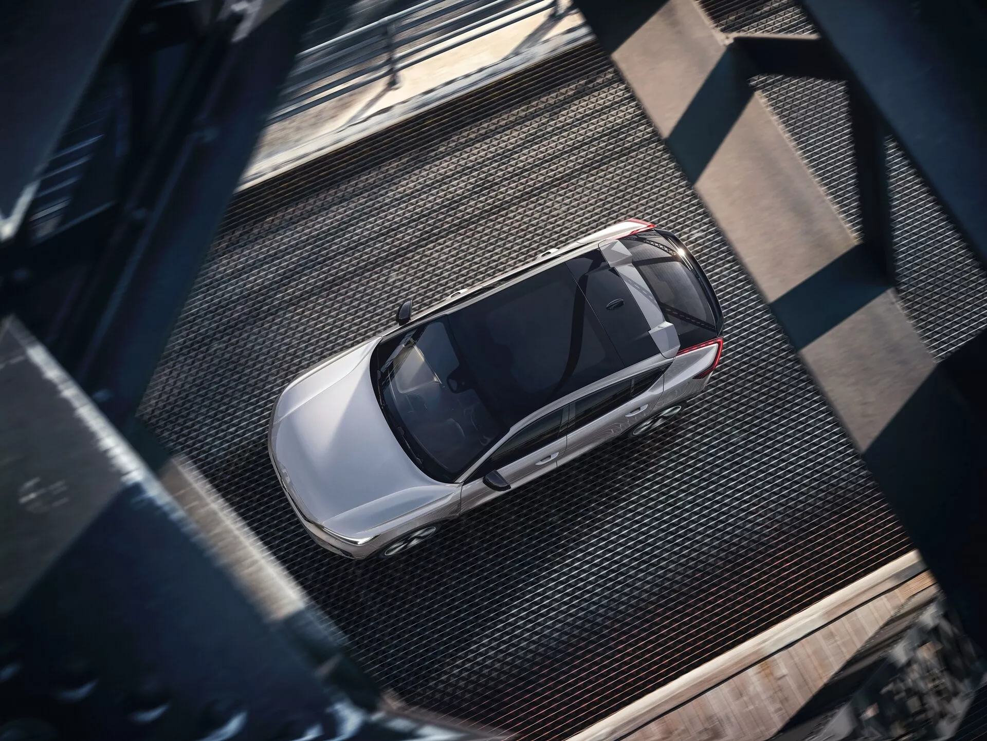 Vista aérea de un Volvo C40 2021 de primera generación, capturado desde una perspectiva elevada. La imagen revela la vista completa del elegante sedán moderno de color metálico y acabado brillante, probablemente plateado o gris claro. Las características visibles incluyen el techo, el capó, el maletero, las ventanas y partes de las puertas, con un prominente techo solar que sugiere una ventilación avanzada. El coche está estacionado sobre una superficie texturizada de estilo industrial con texturas metálicas de rejilla y vigas oscuras remachadas, proyectando sombras que indican una iluminación lateral posiblemente al amanecer o al atardecer.