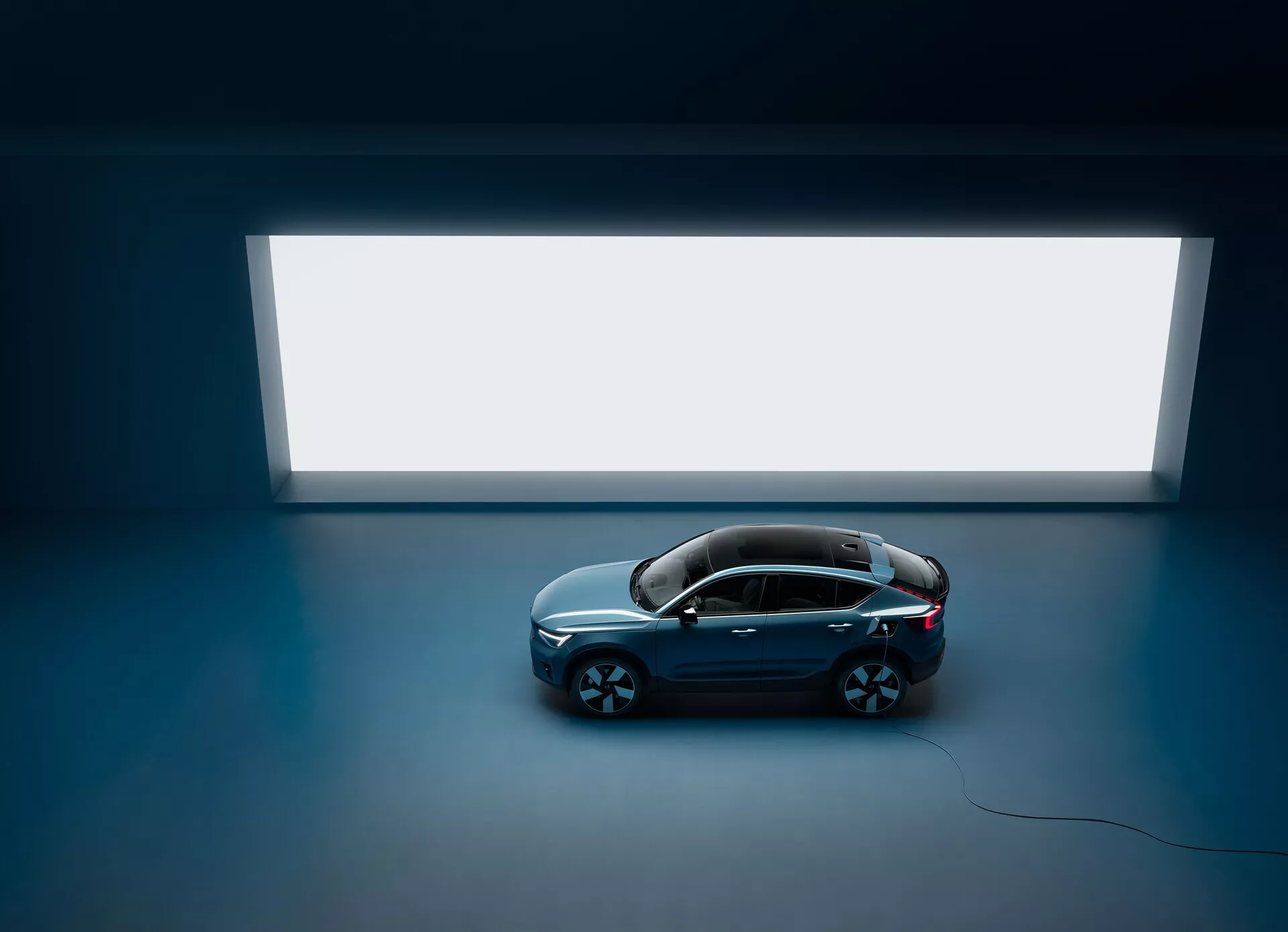 Toma en ángulo alto de un sedán Volvo C40 2021 con cuatro puertas y techo panorámico, visto desde arriba contra un piso oscuro con una fuente de luz rectangular brillante en el fondo, mostrando un diseño limpio y minimalista con una carrocería de color oscuro y acabado brillante y grandes ruedas de aleación de múltiples radios, resaltando su estética lujosa y moderna.