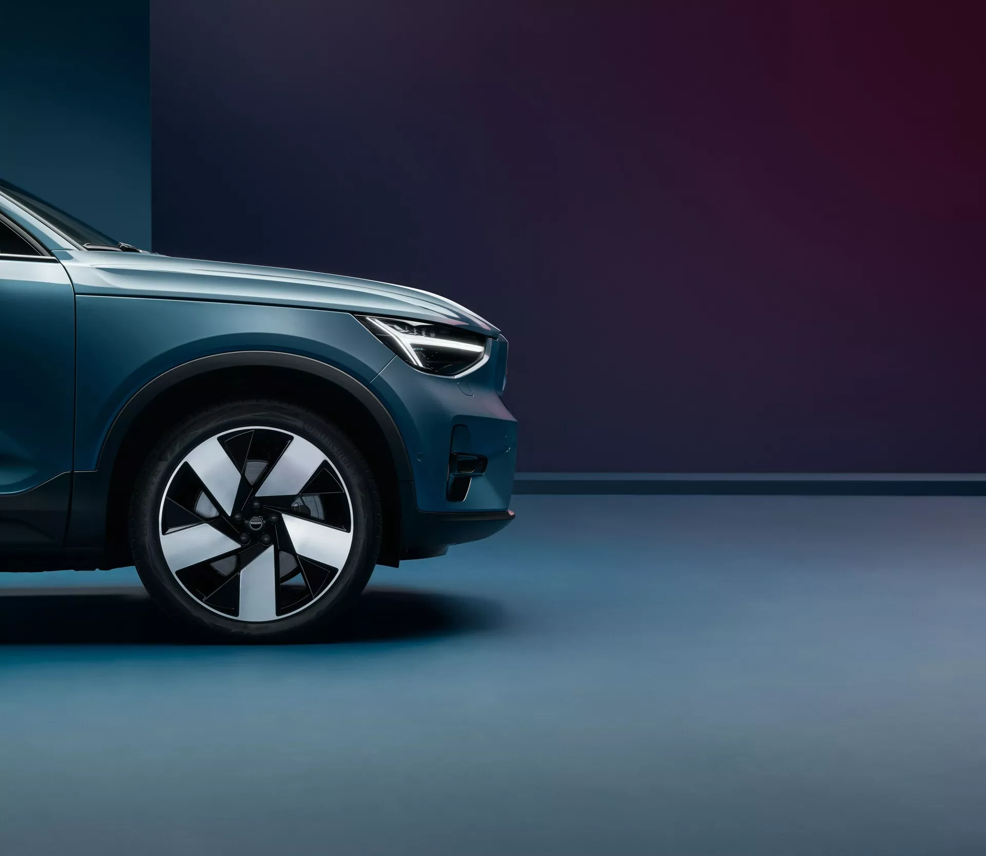 Toma en ángulo bajo del Volvo C40 2021 (primera generación), enfocada en el cuarto delantero desde la rueda delantera hasta el punto medio del capó. Destacan el diseño moderno de los faros, la rueda de aleación de múltiples radios, la pintura metálica brillante y las líneas aerodinámicas. El fondo de doble tono con iluminación suave acentúa el acabado premium y la postura imponente del vehículo.