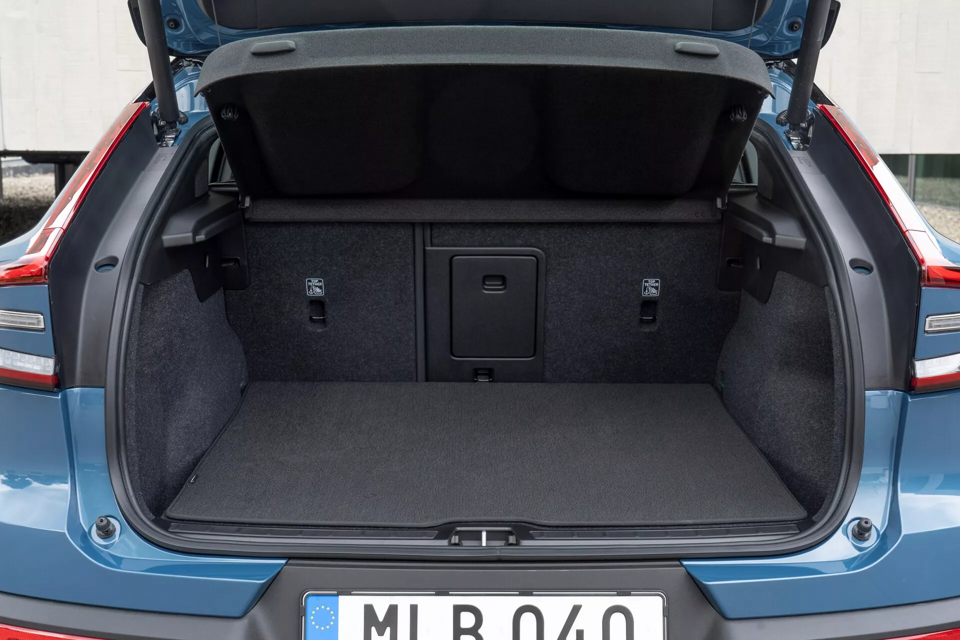 Espacio de carga trasero de un coche moderno, mostrando el maletero del Volvo C40 2021 (1ª generación) desde una perspectiva trasera, ligeramente por debajo de la cintura. La tapa del maletero está abierta, destacando el acolchado estándar del revestimiento interior. El maletero está vacío, mostrando el interior alfombrado, las áreas laterales empotradas y una manija en el medio del piso para acceder a un compartimento de almacenamiento. No se ven ganchos de carga ni puntos de amarre. La matrícula de estilo europeo sugiere un origen europeo.