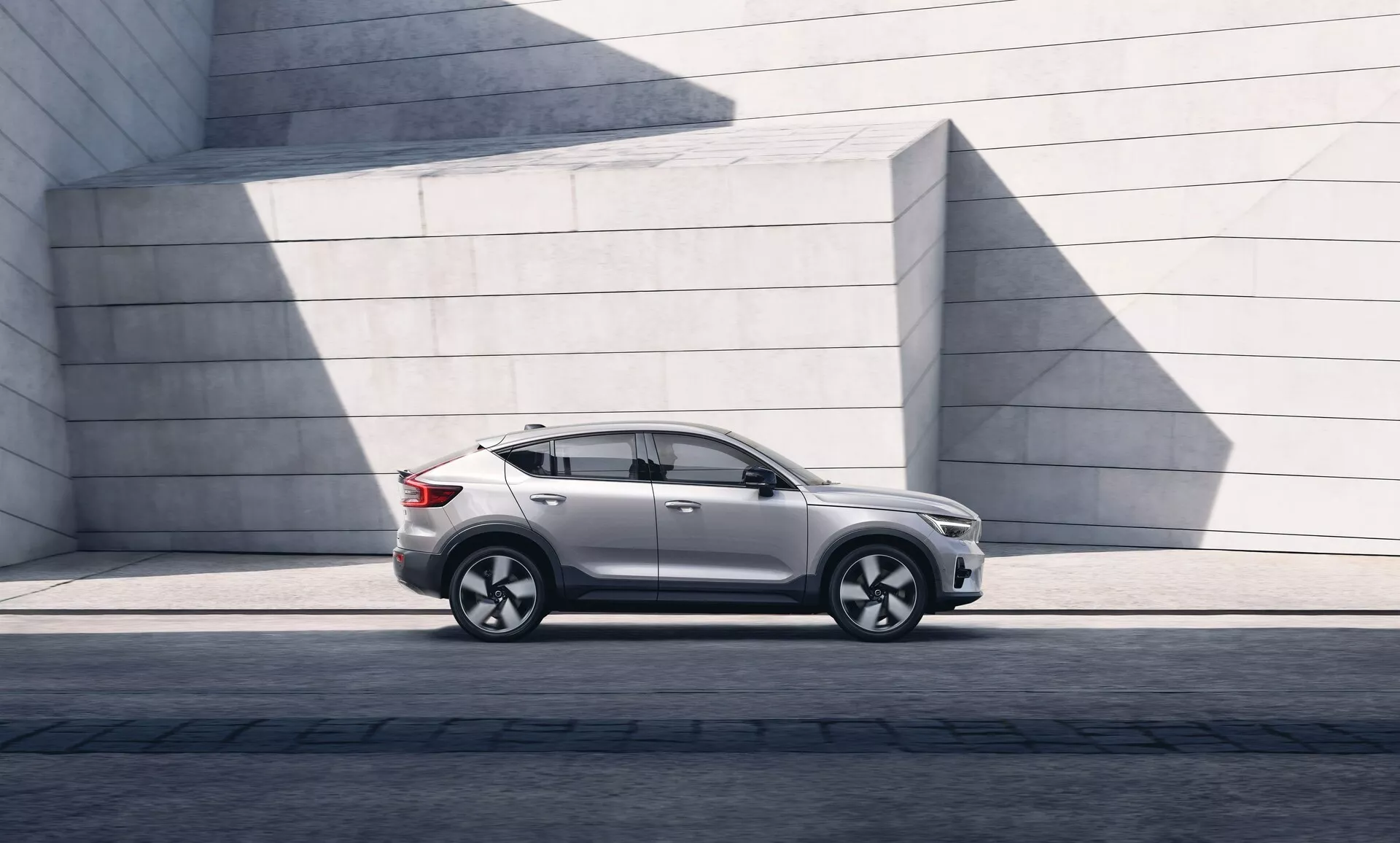 Imagen de un moderno SUV Volvo C40 2021 estacionado en paralelo, mostrando el perfil lateral izquierdo desde el faro delantero izquierdo hasta la luz trasera izquierda. El ángulo resalta la distancia al suelo y la postura sobre una superficie pavimentada. La luz solar crea sombras en el acabado de pintura brillante. El SUV presenta un diseño elegante con una línea de techo suavemente inclinada, contornos distintos en las puertas, ventanas tintadas y espejos laterales contemporáneos. Las ruedas de aleación con un diseño de múltiples radios y neumáticos de perfil bajo indican rendimiento y lujo. Se observan el faldón lateral, los molduras de los arcos de rueda y una línea de carácter a través de las manillas de las puertas, sugiriendo robustez. La ausencia de un escape visible insinúa una posible motorización eléctrica o híbrida. La estructura de fondo de concreto minimalista contrasta con las curvas refinadas del coche, destacando su atractivo urbano y de diseño.