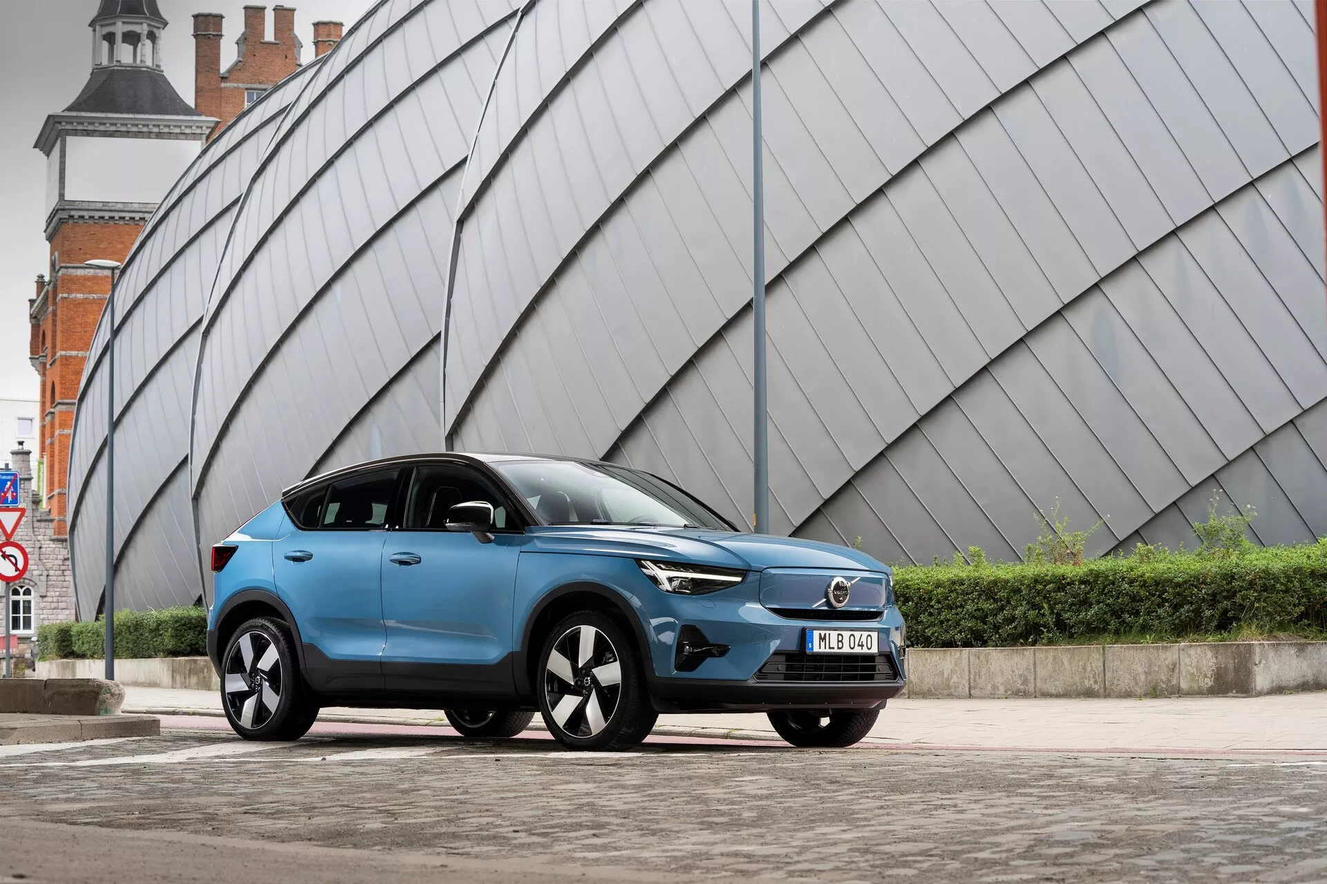 Un moderno SUV Volvo C40 2021 azul estacionado en una área urbana pavimentada con una estructura metálica angular en el fondo. La toma desde un ángulo bajo hacia el lado delantero izquierdo muestra la parrilla frontal del vehículo, los faros LED, el perfil aerodinámico, las llantas de aleación de múltiples radios, los espejos laterales con señales de giro integradas, el acabado de pintura mate o satinada, los vidrios polarizados, la placa de matrícula delantera y las líneas del cuerpo esculpidas.