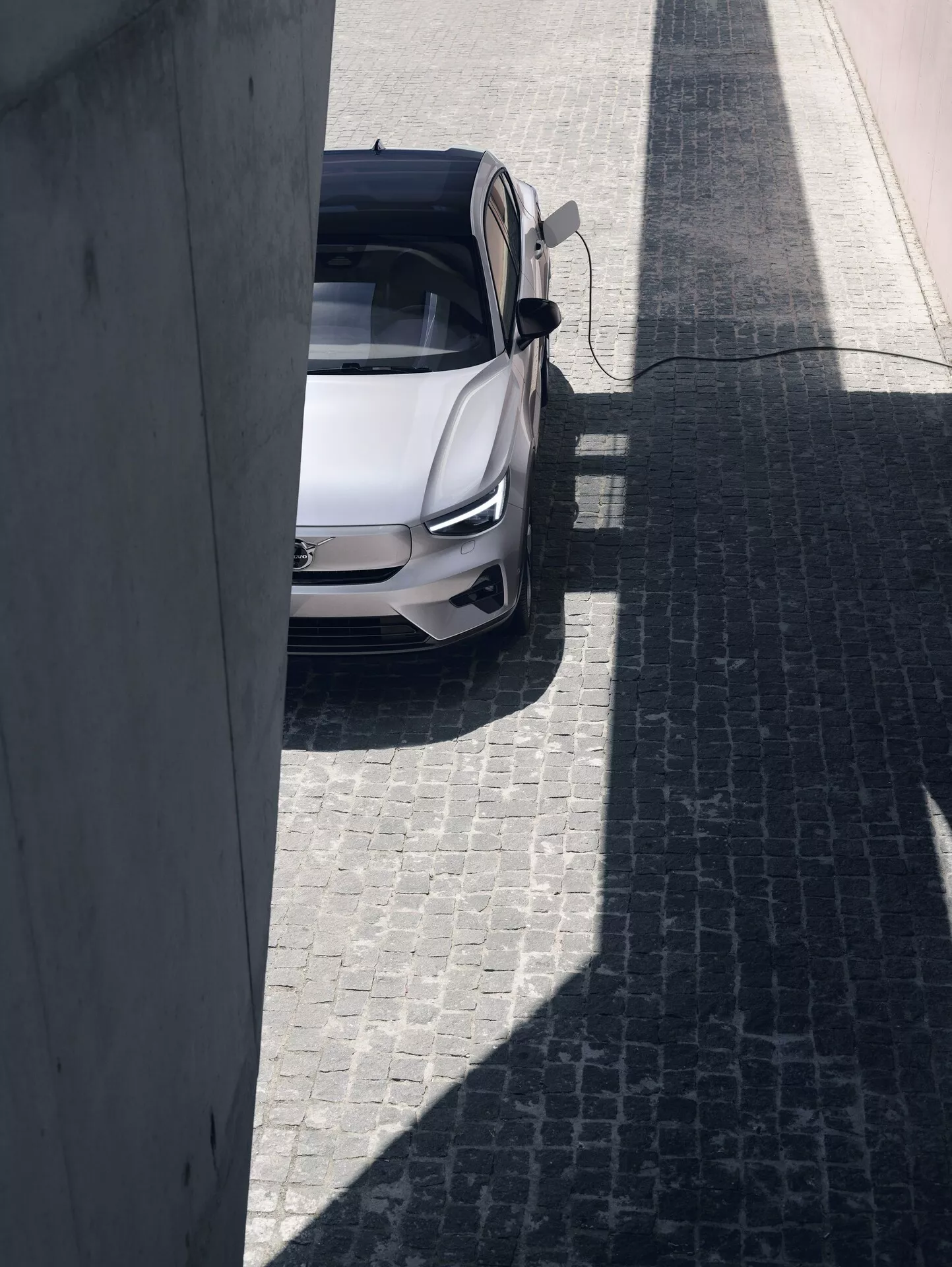 Vista en ángulo alto de un Volvo C40 2021 de primera generación, capturado en un día soleado desde una perspectiva elevada. El coche está centrado en una calle adoquinada, con un color claro, posiblemente blanco o plateado, y un techo solar panorámico. La puerta del lado del conductor está abierta, proporcionando una visión del interior, destacando el diseño moderno y deportivo del sedán con potenciales características avanzadas de automoción.