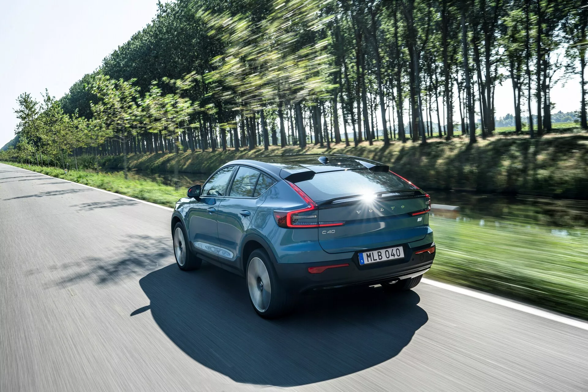 Toma dinámica del Volvo C40 2021 (1ª generación) en movimiento en un día soleado, vista trasera de tres cuartos que muestra el perfil aerodinámico del vehículo. El coche, pintado en un azul brillante, presenta distintivas luces traseras LED, ruedas borrosas por el movimiento y árboles reflejando su velocidad. Detalles destacables incluyen un techo panorámico, spoiler trasero y placa de matrícula en el portón trasero, resaltando elementos de diseño moderno y tecnología avanzada de iluminación.