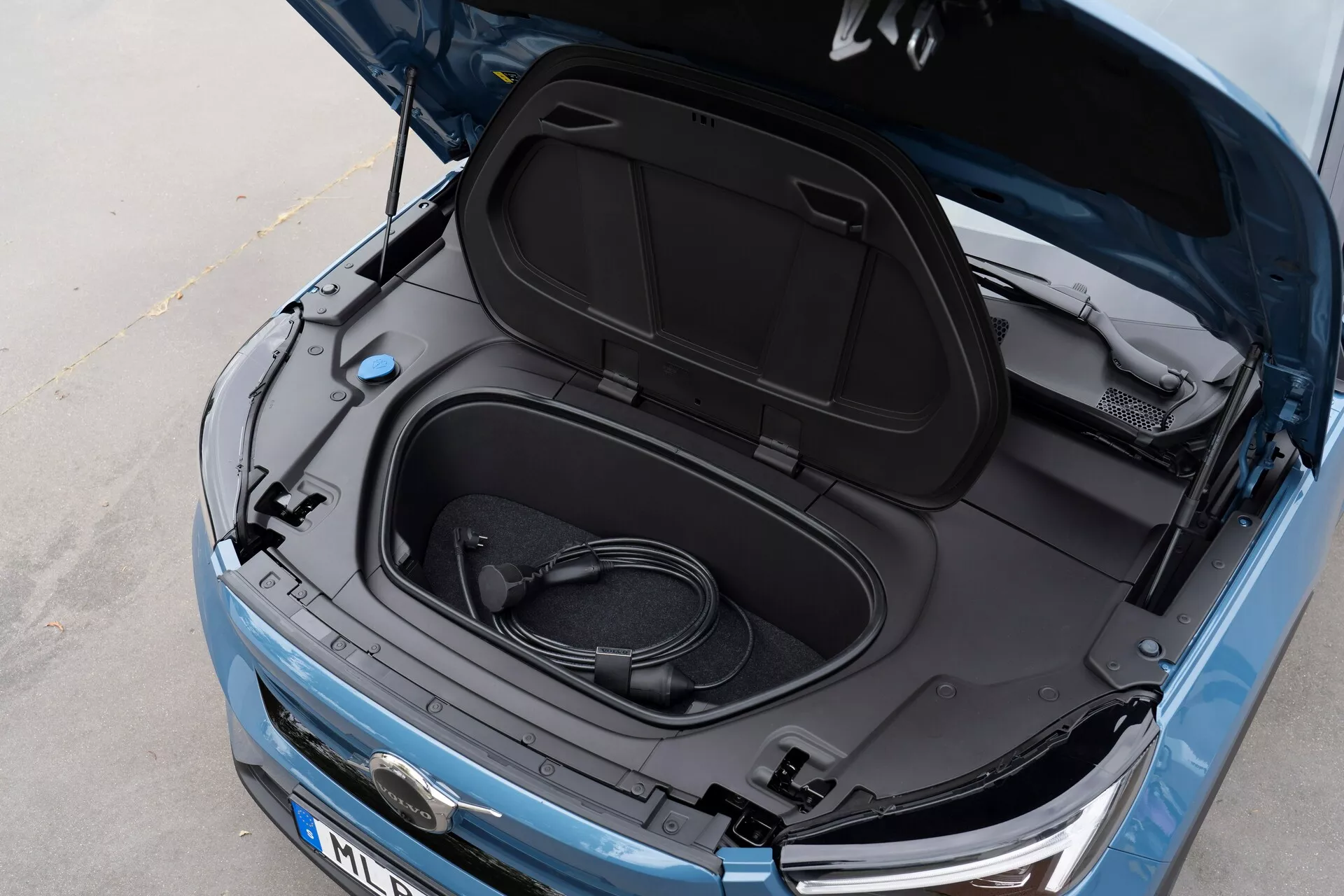 Vista en primer plano desde arriba en ángulo del Volvo C40 2021 (1ª generación) con el frunk abierto, revelando espacio de almacenamiento vacío y un cable de carga enrollado, indicando que es un vehículo eléctrico. Pintura azul metálica, interior de la tapa del frunk negro con etiquetas informativas, vista parcial del logotipo del fabricante, faros elegantes y rejilla frontal visibles, sugiriendo un EV con tecnología LED moderna y estética aerodinámica.