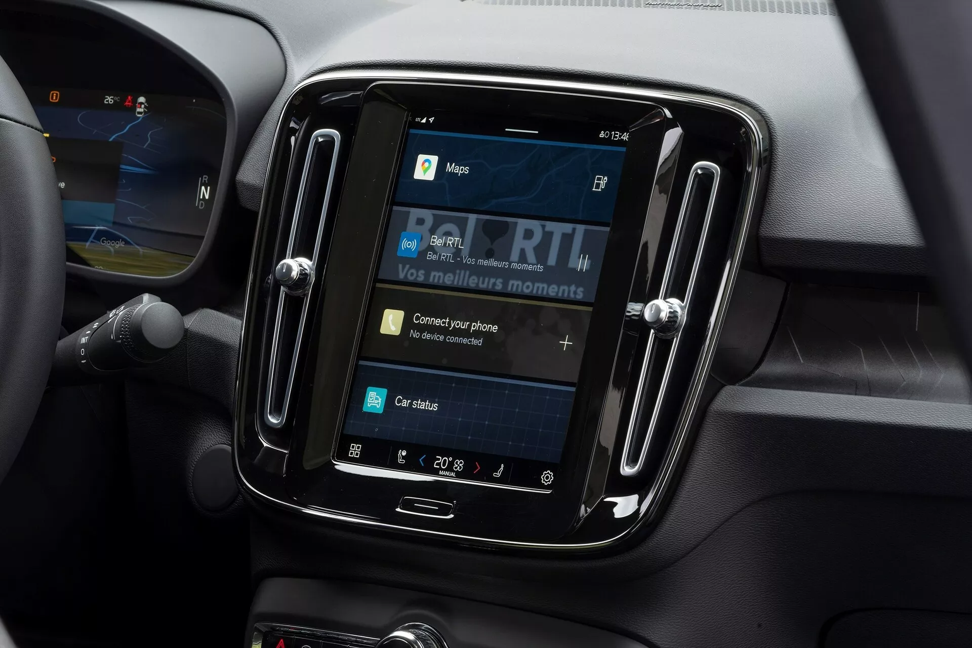 Vista de cerca del sistema de infoentretenimiento dentro de un Volvo C40 2021. La toma, hecha desde un ángulo levemente hacia abajo, se centra en la pantalla táctil enmarcada en un bisel negro brillante. La pantalla muestra opciones como mapas, medios, conectividad del teléfono y estado del coche, con 'Conecta tu teléfono' como un elemento interactivo. Alrededor del sistema de infoentretenimiento se ven materiales suaves al tacto y líneas de contorno que indican ventilaciones de aire, aludiendo a un interior de coche moderno. La temperatura se muestra en 20° Celsius, y una parte del volante es visible a la izquierda.