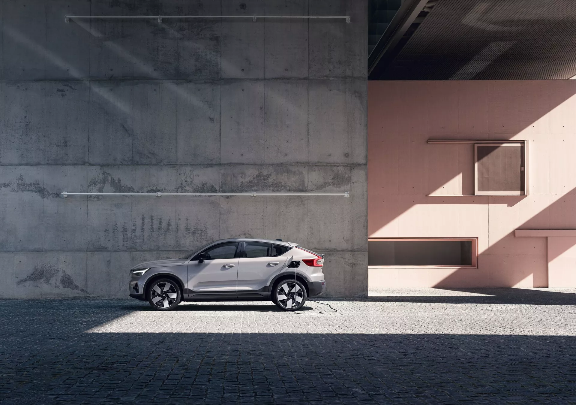 Perfil lateral del moderno SUV Volvo C40 2021 (1ª Generación) contra un fondo urbano con paredes de concreto y color melocotón. El coche presenta una pintura blanca reflectante, llamativas llantas de aleación, ventanas tintadas, rieles de techo plateados y señales de giro integradas en los espejos retrovisores. La foto resalta las líneas y curvas del diseño del coche bajo la luz natural del día, añadiendo un sentido de lujo y sofisticación.