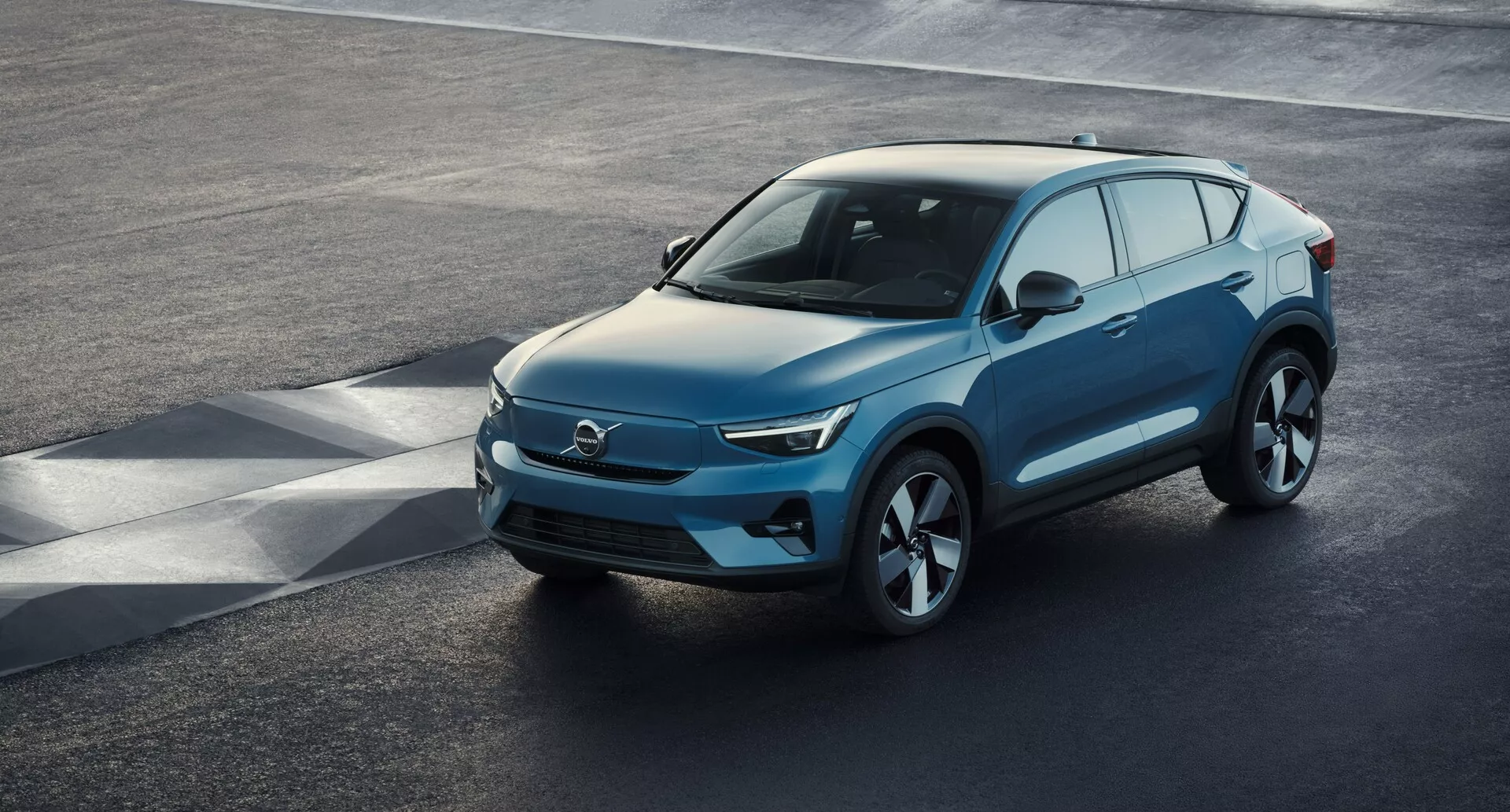 Vista de ángulo elevado de un SUV moderno Volvo C40 2021 (1ª generación) estacionado en una superficie de asfalto, mostrando una perspectiva frontal de tres cuartos a alta altura. La elegante área frontal, probablemente característica de un vehículo eléctrico con mínimas tomas de aire, se combina con faros LED nítidos, un capó suave en pendiente y un perfil lateral contorneado con grandes ruedas de aleación. Espejos laterales con señal de giro integrada, amplio espacio de ventanas con posible tinte de privacidad y una línea de techo suavemente inclinada con barras de techo para utilidad son características notables. La vista clara captura el diseño lujoso del vehículo, avanzada aerodinámica y elementos tecnológicos modernos.
