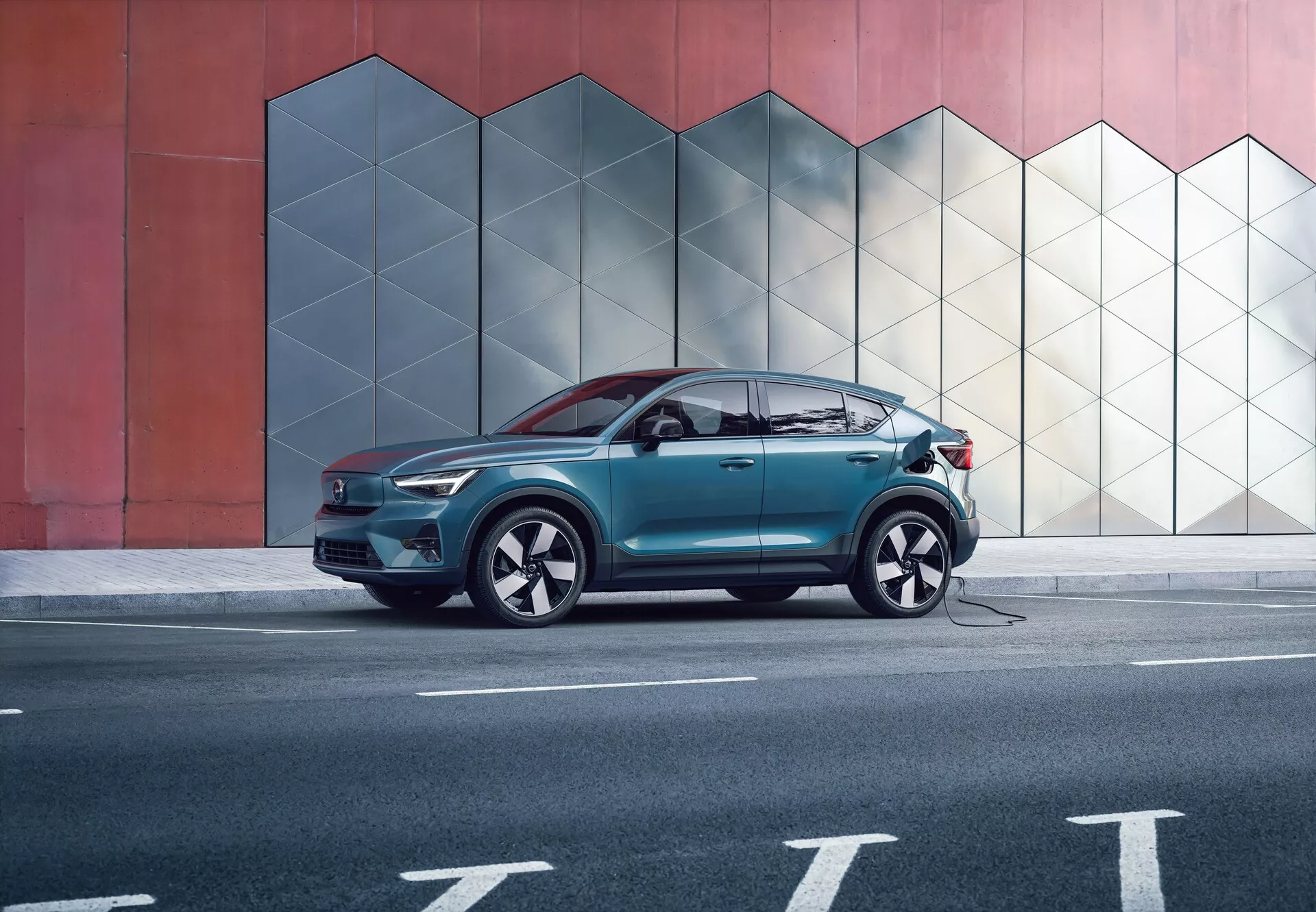 Vista de perfil lateral del SUV moderno Volvo C40 del 2021 (1ª generación) frente a una pared urbana con un patrón geométrico, capturada desde una distancia media. La imagen resalta las líneas aerodinámicas del cuerpo, las ventanas con acabado cromado y las grandes ruedas con neumáticos de perfil bajo. El SUV tiene un cable de carga conectado en su lateral, indicando tecnología de vehículo eléctrico. No hay personas presentes, lo que enfatiza el diseño y las características del coche.
