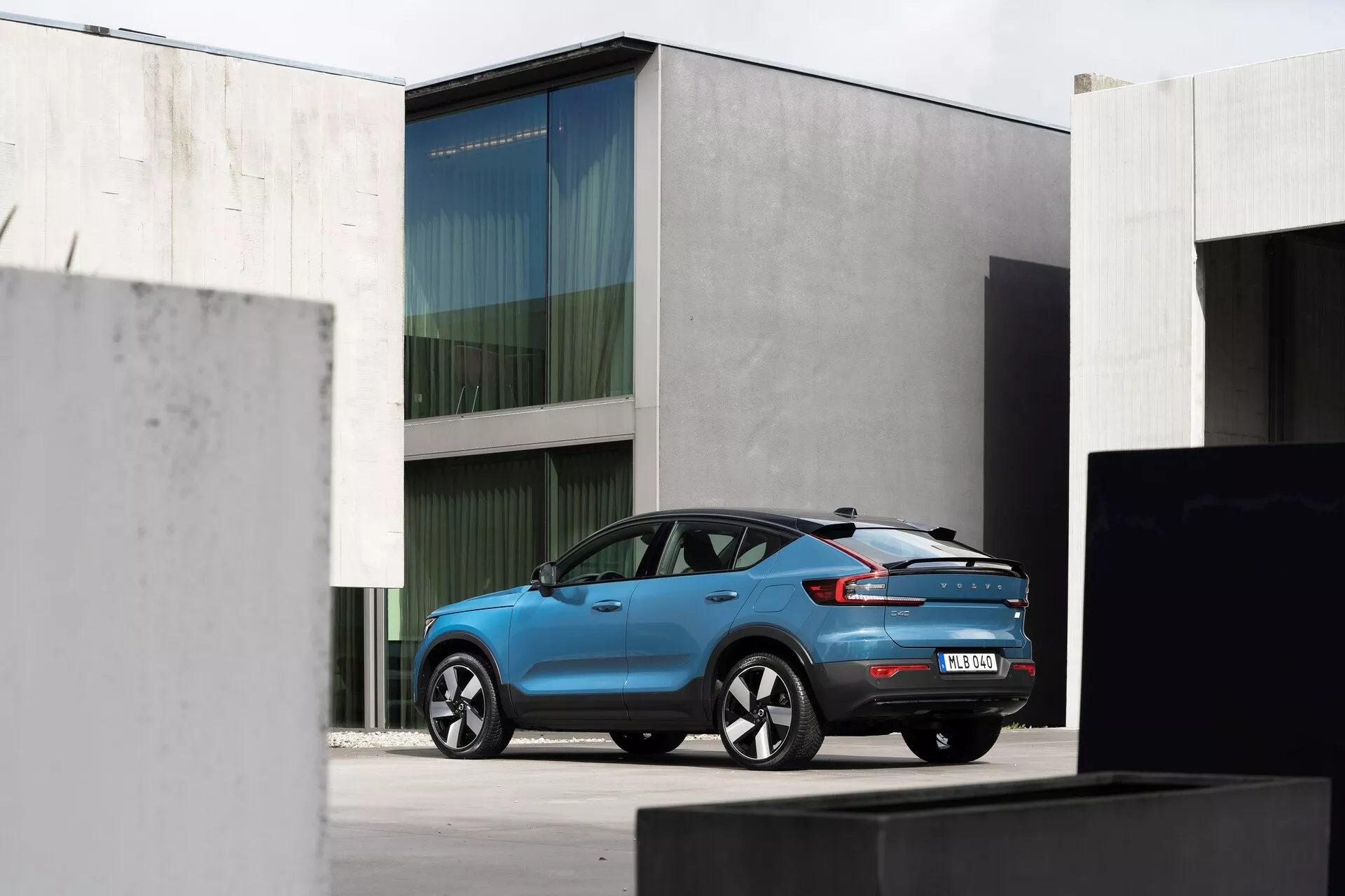 Vista trasera de tres cuartos del Volvo C40 2021 (1ª generación) SUV compacto en color azul brillante, mostrado en un entorno urbano con arquitectura minimalista. El coche presenta luces traseras LED alargadas, un alerón trasero integrado, una antena de aleta de tiburón, llantas de aleación con un distintivo diseño de radios y sensores en el parachoques trasero. La iluminación natural y difusa resalta su diseño moderno.