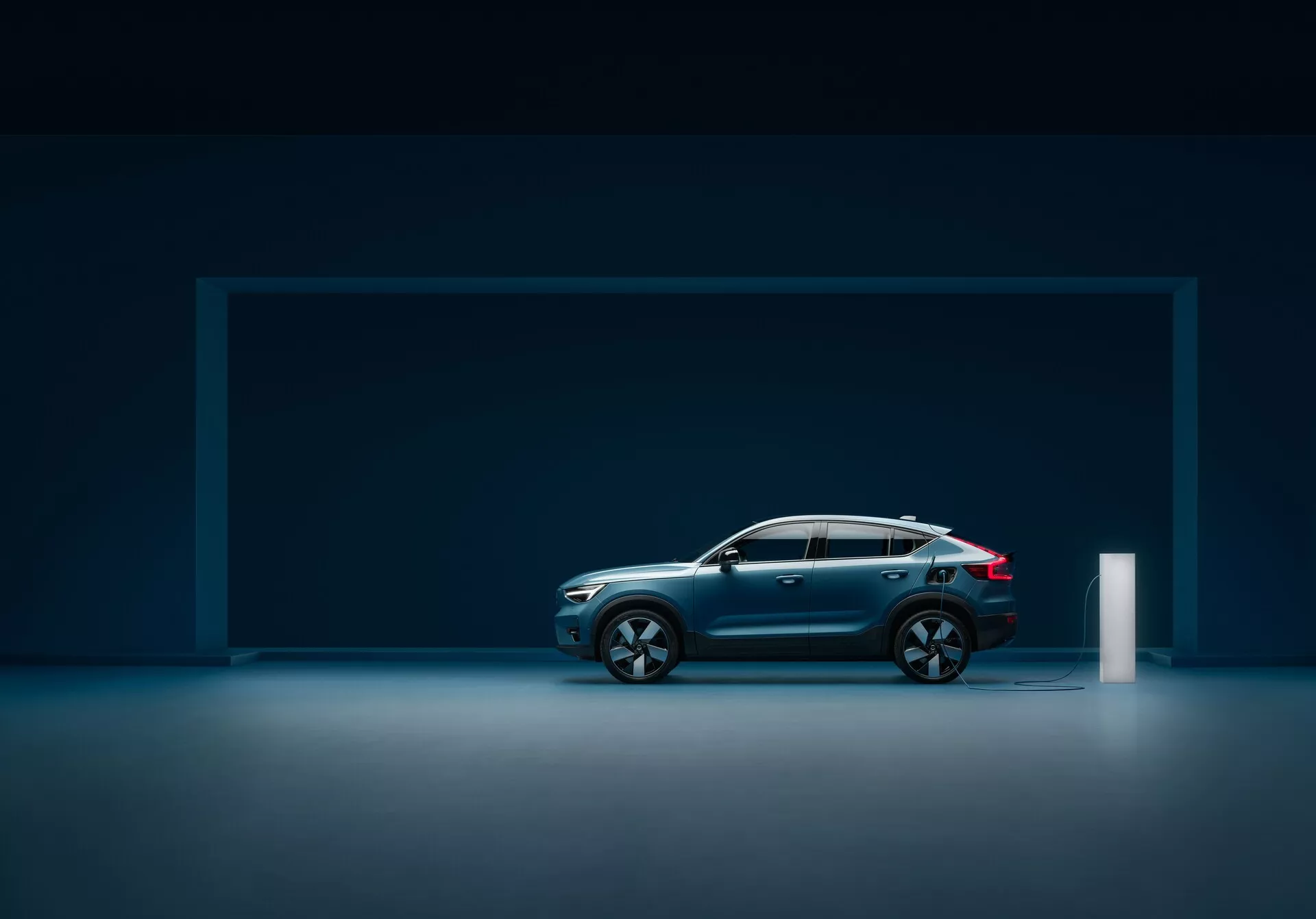 Vista de perfil lateral de un moderno coche Volvo C40 2021 (primera generación) en un estudio fotográfico con suelo reflectante, mostrando un diseño elegante, pintura metálica azul oscuro, línea de techo inclinada, grandes llantas de aleación multirradio, líneas contorneadas, construcción robusta, espejos laterales integrados con intermitentes, luces traseras LED y puerto de carga eléctrica.