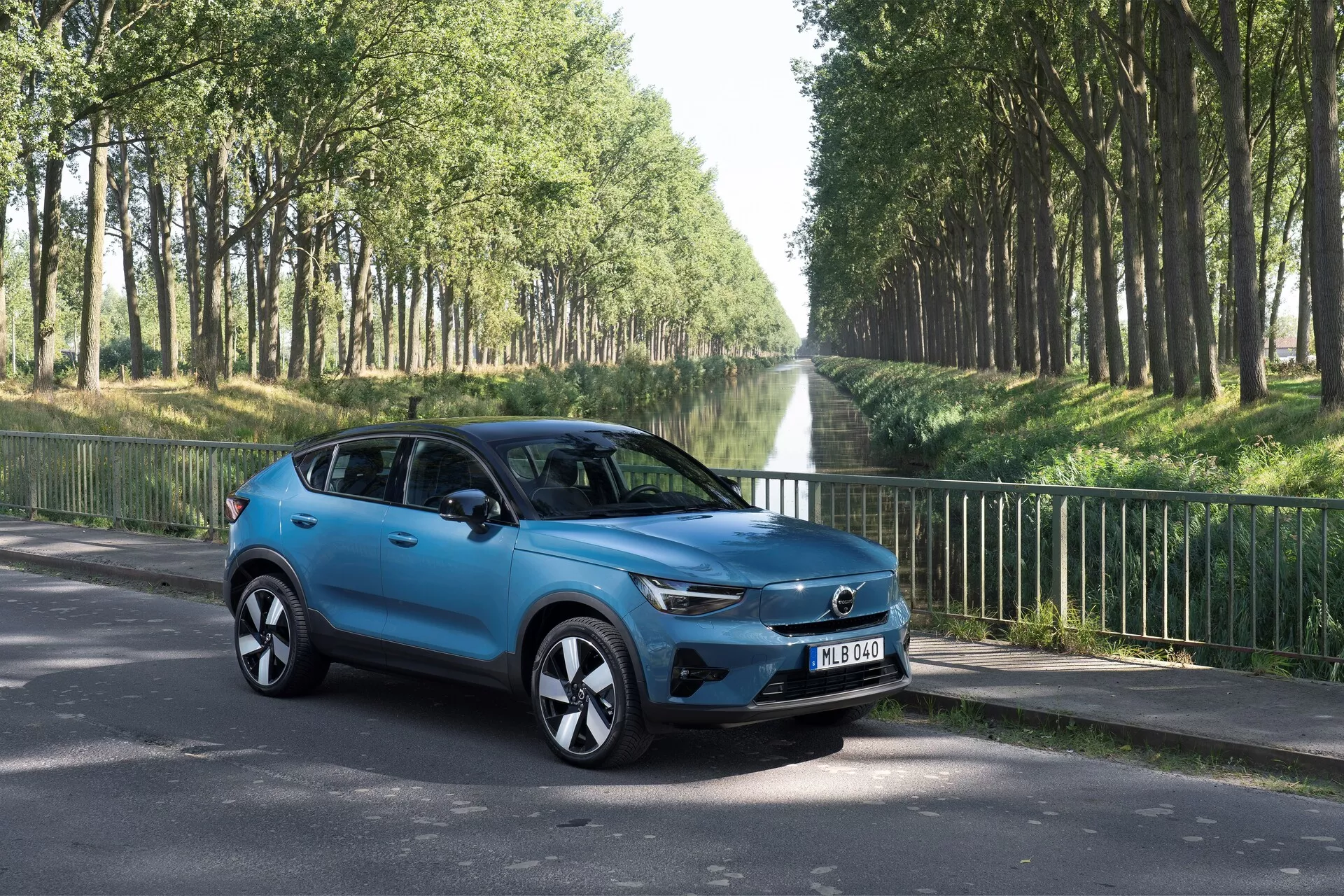 Volvo C40 2021 (Primera Generación) SUV compacto en vista de tres cuartos desde el frente izquierdo en una carretera asfaltada. Muestra aspectos de diseño moderno como la parrilla prominente con el emblema centrado, faros LED elegantes, llantas de aleación estilizadas, espejos laterales con señales de giro integradas, carrocería aerodinámica esculpida con un acabado de pintura azul brillante, ventanas tintadas y cuatro puertas. Ambientado en un entorno al aire libre con árboles bordeando la carretera, centrado en el atractivo estético y las características tecnológicas del vehículo.
