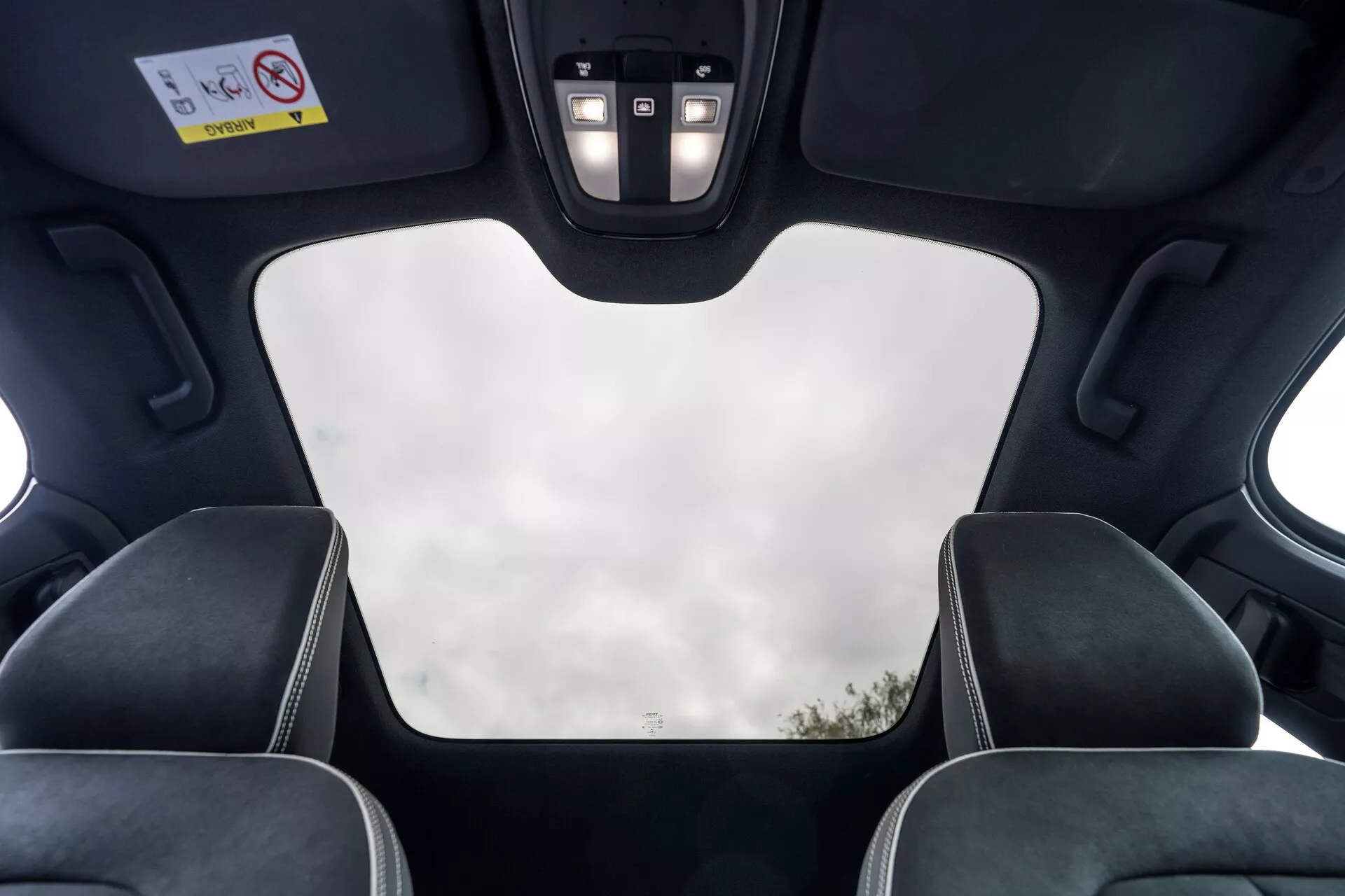 Vista interior del Volvo C40 2021 (1ª generación) desde la perspectiva del pasajero trasero, mostrando características del techo como un techo panorámico cerrado con vista despejada del cielo, revestimiento interior negro, huecos del mecanismo del techo, luces LED y panel de control con botones para varias funciones. La imagen también resalta las cabeceras de los asientos delanteros con costuras contrastantes, enfatizando el diseño moderno y lujoso del vehículo y sus características de alta gama.