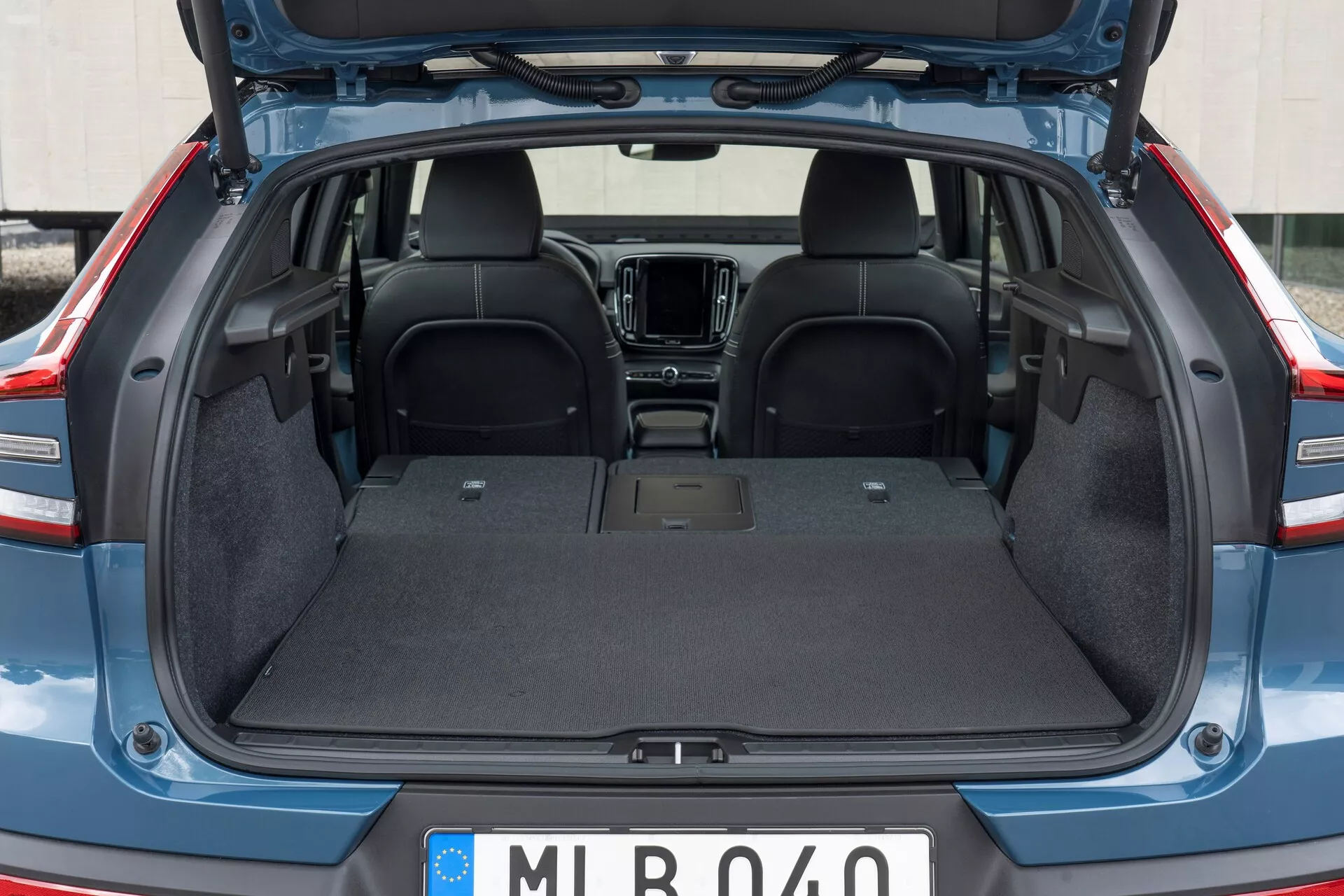 Interior trasero del Volvo C40 2021 (1ª generación) con el maletero abierto, mostrando el área de carga vacía con forro de alfombra duradera, superficie plana y sin intrusiones visibles. La parte superior de los asientos traseros hechos de tela oscura está presente, con una vista parcial de los elementos de la cabina delantera como la consola central y el sistema de infoentretenimiento. La tapa del maletero está levantada, revelando los bordes con moldura de plástico/composite.