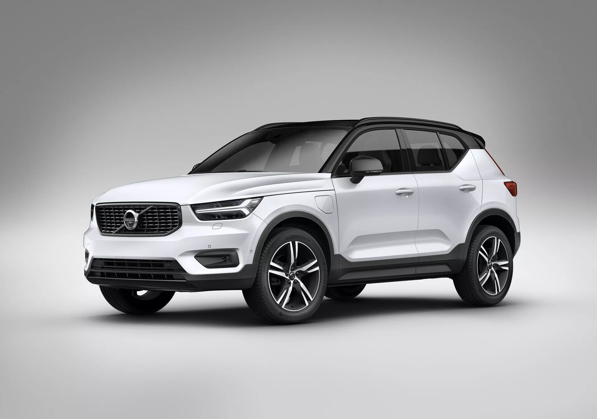 Fotografía de estudio de un SUV moderno Volvo XC40 2018 (1ª generación) en color blanco. Tomada desde un ángulo bajo, la imagen destaca la postura y los elementos de diseño del coche, incluyendo una vista frontal de tres cuartos que muestra los perfiles delantero y lateral. Los faros tienen una forma moderna, posiblemente con tecnología LED, mientras la parrilla incluye el emblema de la marca y líneas horizontales minimalistas. El parachoques inferior integra luces antiniebla o entradas de aire, insinuando tecnología avanzada. Destacan las grandes llantas de aleación, posiblemente de 18 pulgadas, manillas de las puertas y tapas de espejos en color de la carrocería, y techo panorámico. La línea del techo se estrecha hacia la parte trasera, acentuando el aspecto de lujo con ribetes cromados o metálicos alrededor de las ventanas. El acabado de pintura suave y brillante y el fondo de estudio resaltan las características del vehículo sin distracciones, sugiriendo un enfoque en seguridad, comodidad, entretenimiento e innovadores sistemas de propulsión.