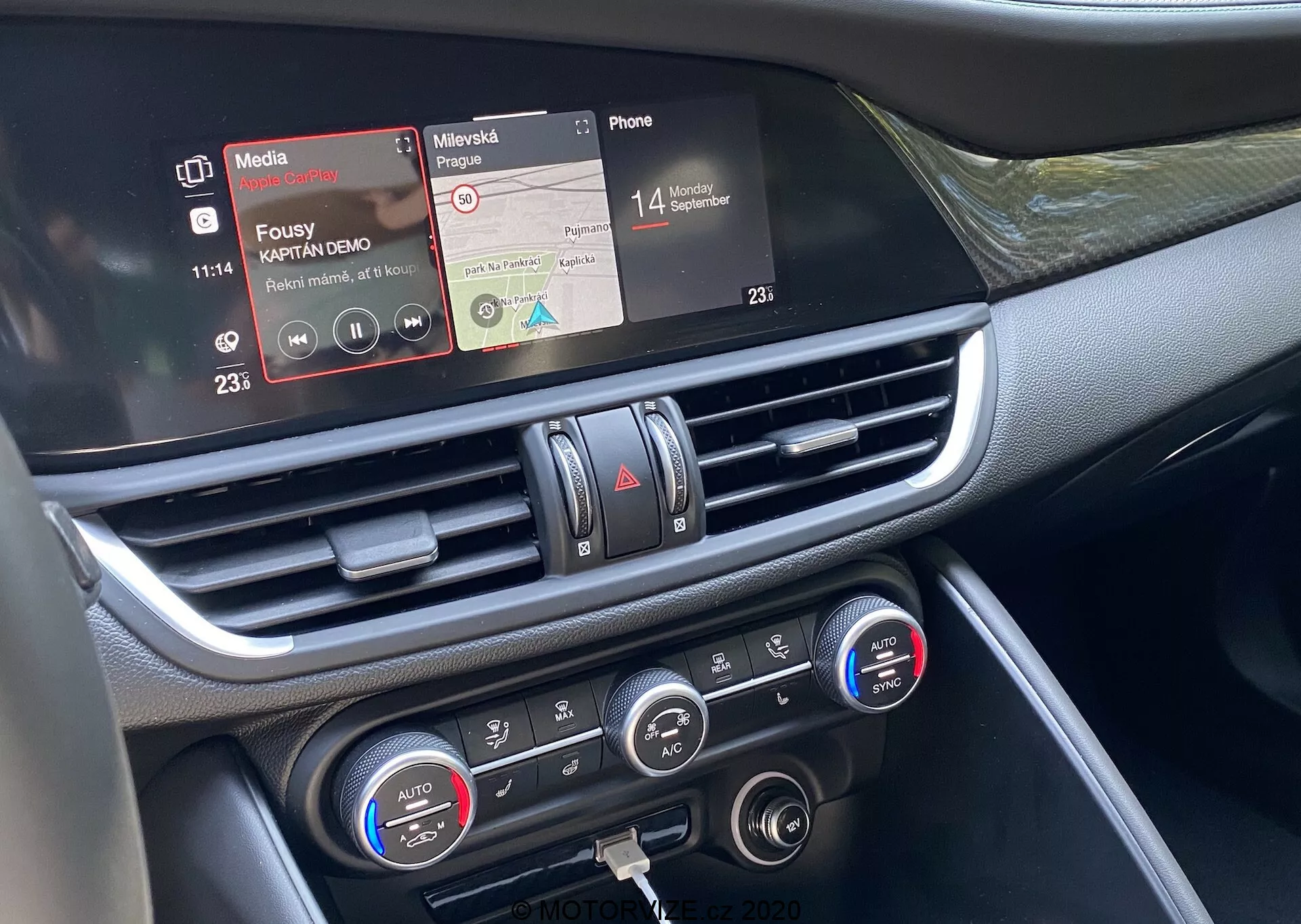 Vista de cerca de la sección central del tablero del Alfa Romeo Giulia 2016 desde el asiento del pasajero delantero. La imagen en primer plano destaca la pantalla táctil de infoentretenimiento con funciones de reproducción de medios y conectividad telefónica, la salida de aire central con detalles cromados y los controles de clima con ajustes de temperatura de zona dual y pantallas digitales. Los materiales del tablero parecen incluir plásticos y detalles metálicos o cromados, mostrando la avanzada tecnología interior del vehículo centrada en la conectividad, reproducción de medios y control de clima.