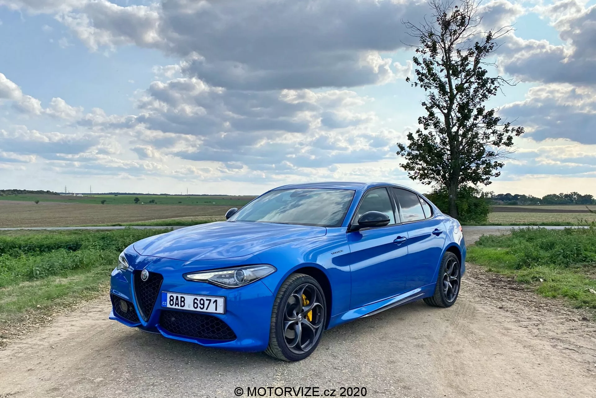 Imagen de un Alfa Romeo Giulia 2016 sedán deportivo azul brillante estacionado en un camino de grava, con un cielo parcialmente nublado de fondo. El coche se captura desde un ángulo delantero de tres cuartos, mostrando su distintiva parrilla frontal 'Trilobo', pinzas de freno amarillas, grandes llantas de aleación multirradio, faros LED elegantes y molduras cromadas en las ventanas. El vehículo está ligeramente descentrado hacia la izquierda con la rueda delantera izquierda apuntando hacia afuera, lo que le da una postura dinámica.