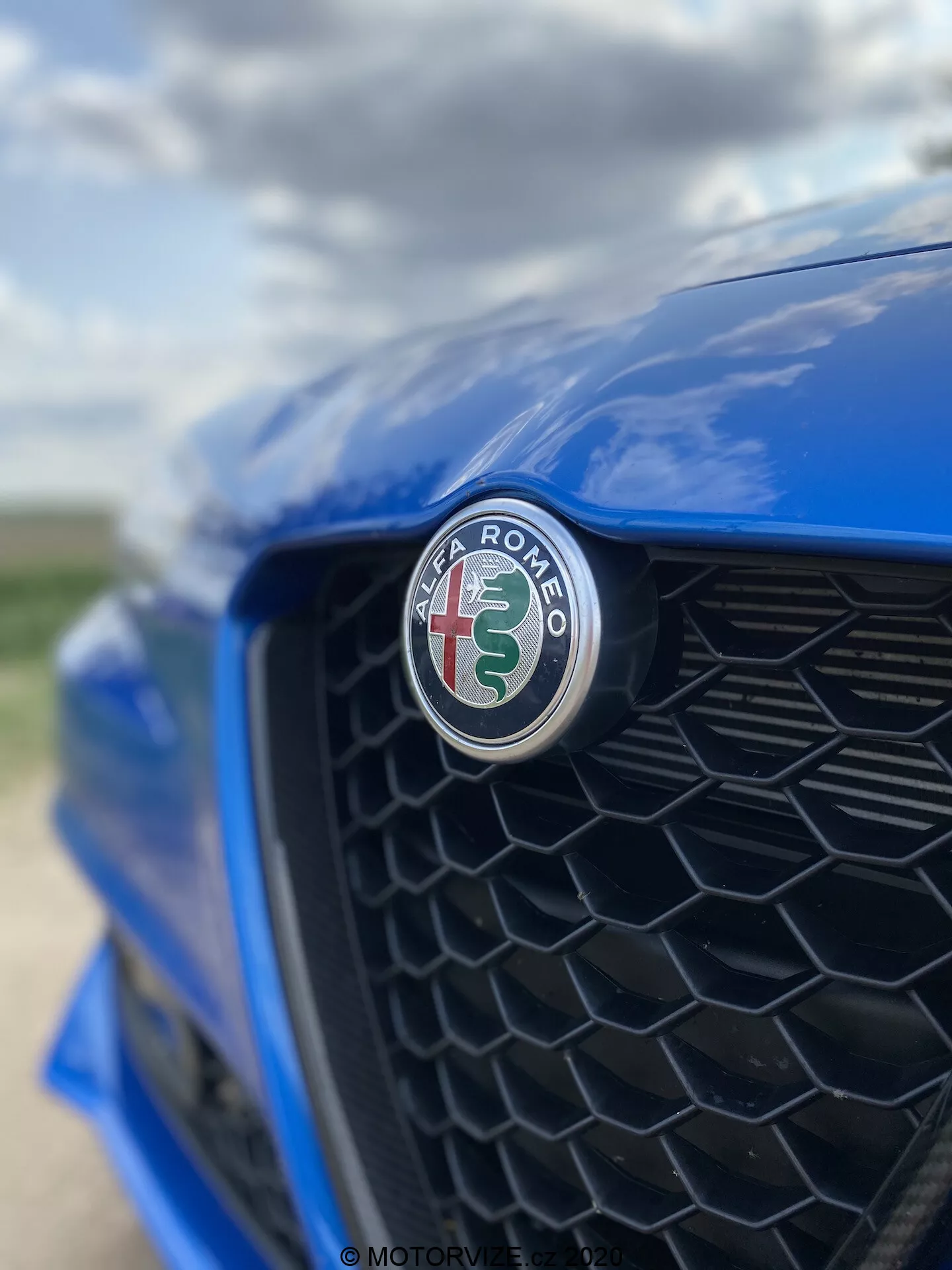Primer plano de la sección frontal de un Alfa Romeo Giulia 2016 azul, centrándose en el emblema del fabricante con una cruz roja y una serpiente verde sobre un fondo blanco. Tomada desde un ángulo descentrado hacia el lado del conductor, la rejilla de panal negro está suavemente desenfocada, resaltando el enfoque nítido del emblema y el acabado brillante.