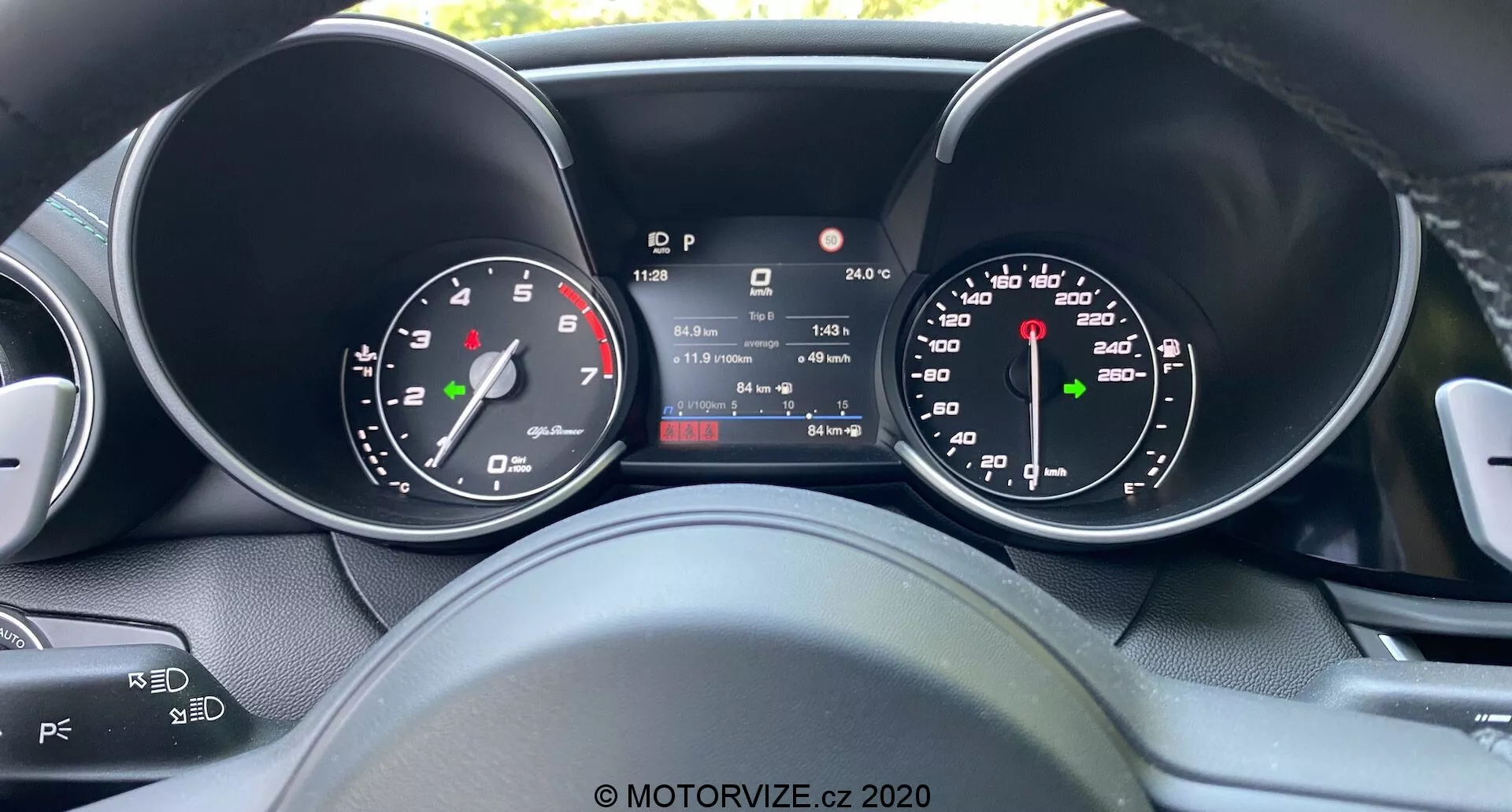 Vista desde el conductor del cuadro de instrumentos del Alfa Romeo Giulia 2016 con pantalla digital central que muestra velocidad, marcha, temperatura y lectura del odómetro, flanqueada por indicadores de velocímetro y tacómetro analógicos, mostrando una combinación de diseño tradicional y tecnología moderna con vistas parciales de los controles del volante para audio y ajustes del control de crucero.