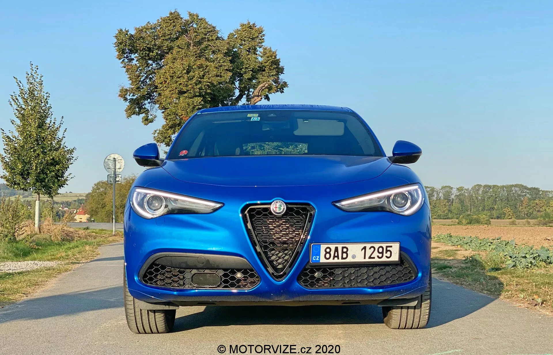 Vista frontal de un Alfa-Romeo Stelvio 2017 azul metálico, mostrando su postura agresiva y deportiva. Las características clave incluyen una parrilla triangular grande y distintiva con el logotipo de la marca, faros elegantes con luces diurnas, grandes tomas de aire en el parachoques delantero y un capó con líneas elevadas. La pintura azul metálica indica un acabado premium.