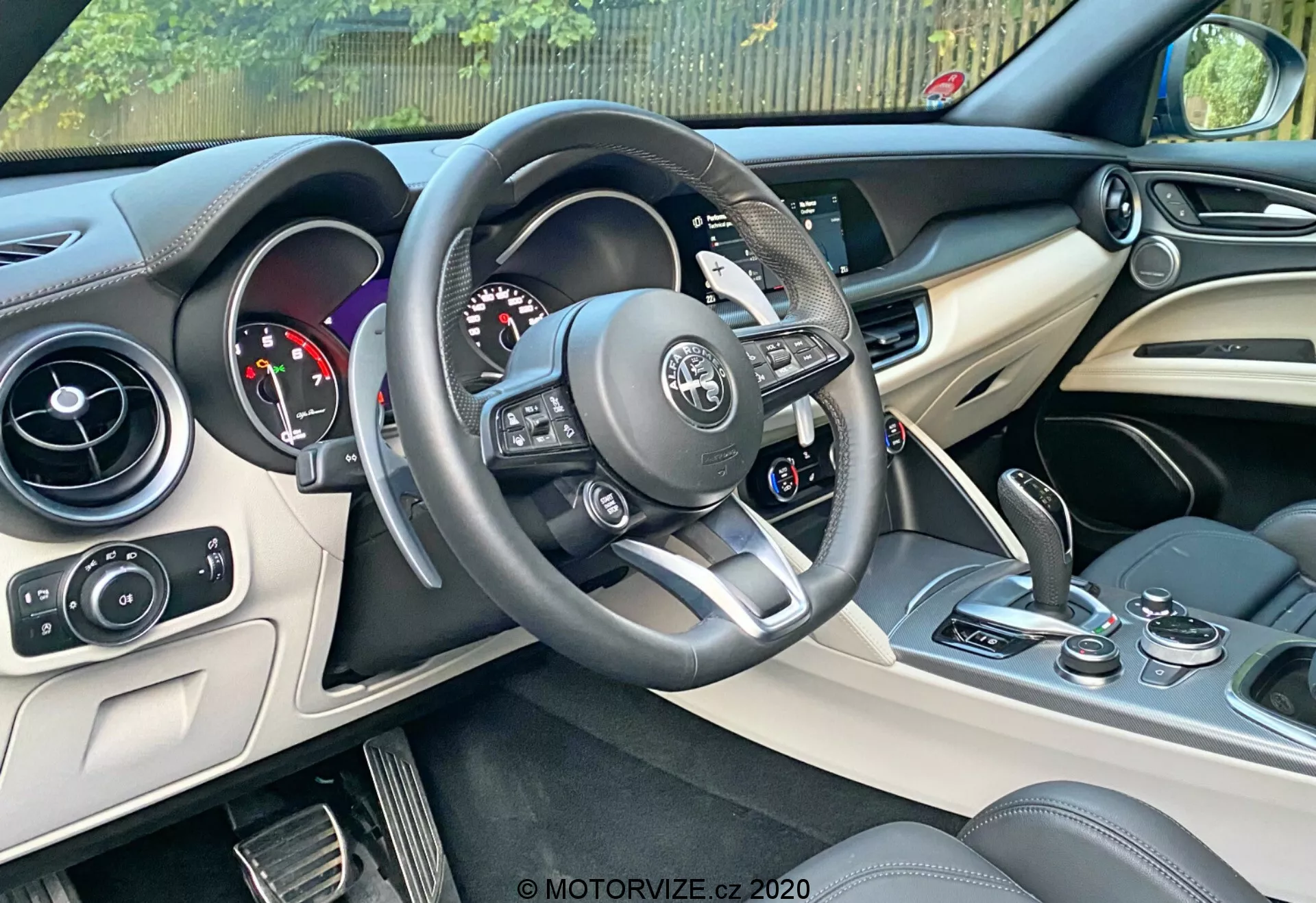 Imagen del interior de un Alfa Romeo Stelvio 2017 enfocada en el área del conductor. Muestra un vehículo con volante a la izquierda, volante multifuncional forrado en cuero con acentos plateados y controles montados, un cuadro de instrumentos con esferas analógicas y pantalla digital, consola central con sistema de infoentretenimiento y controles de clima, y un vistazo a los asientos en diseño bicolor de cuero. Elementos de diseño moderno incluyen detalles en plata o cromo en la consola y materiales lujosos en el tablero y puertas. Incluye botón de encendido/apagado cerca de la consola central.