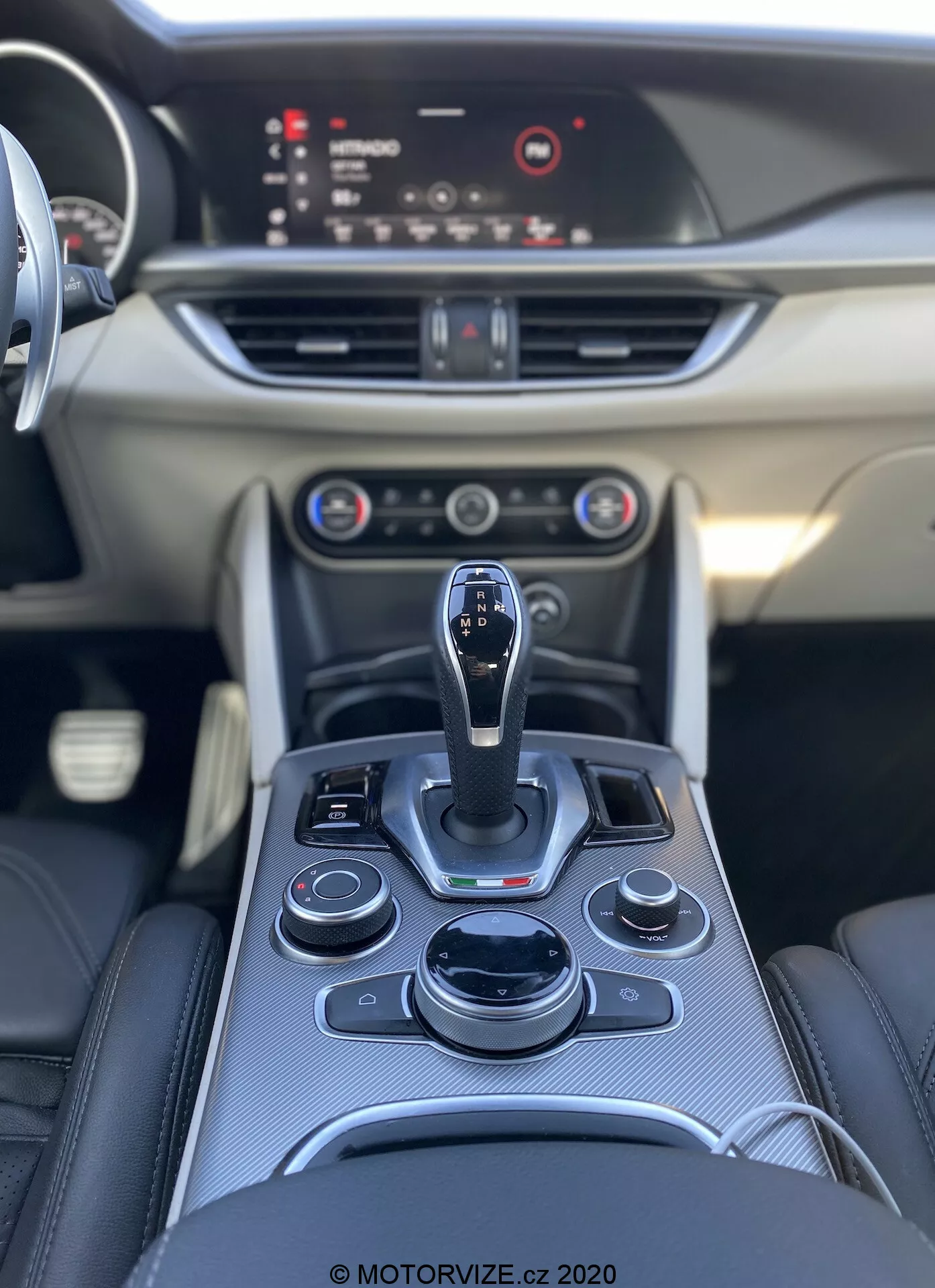 Vista interior del Alfa Romeo Stelvio 2017, mostrando la consola central con una palanca de cambios automática y una disposición ergonómica de botones y diales, incluyendo el sistema de infoentretenimiento, controles de clima y posiblemente modos de conducción. El tablero presenta controles adicionales, salidas de aire y una pantalla de infoentretenimiento destacada. Tablero en el área del pasajero liso con detalles decorativos. Fotografía de alta calidad que destaca los materiales premium y el diseño elegante. © MOTORVIZE.cz