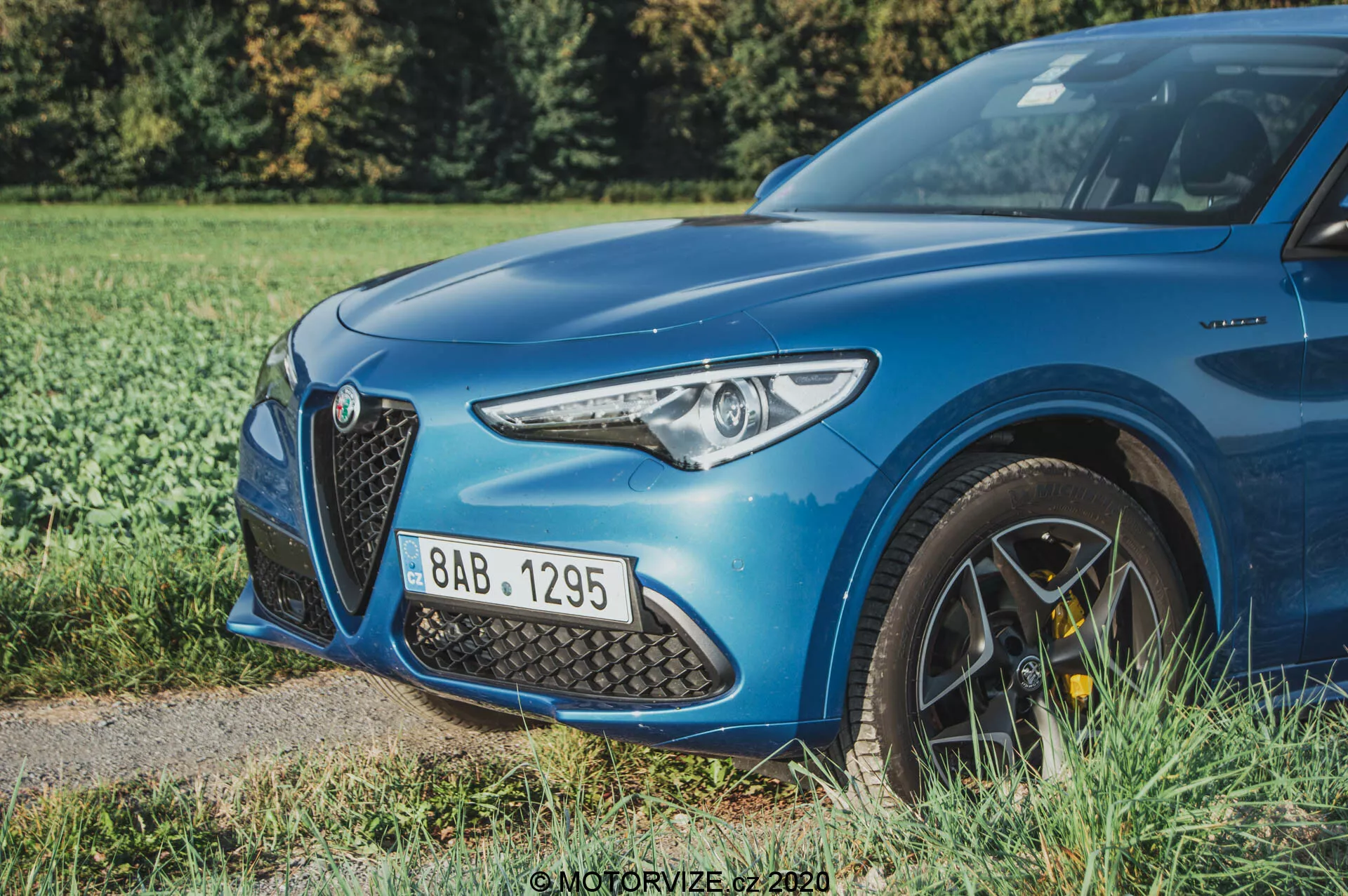 Un Alfa Romeo Stelvio azul de 2017 mostrado desde un ángulo bajo de tres cuartos delanteros, destacando el lado frontal izquierdo incluidas las luces LED modernas, la gran parrilla triangular intrincada, el capó aerodinámicamente perfilado y parte de la llanta de aleación de múltiples radios con pinzas de freno de alto rendimiento amarillas. El coche está estacionado en un borde de césped junto a una superficie pavimentada, con una profundidad de campo somera que desenfoca el fondo de vegetación, enfatizando los detalles nítidos del diseño y las características deportivas del coche.