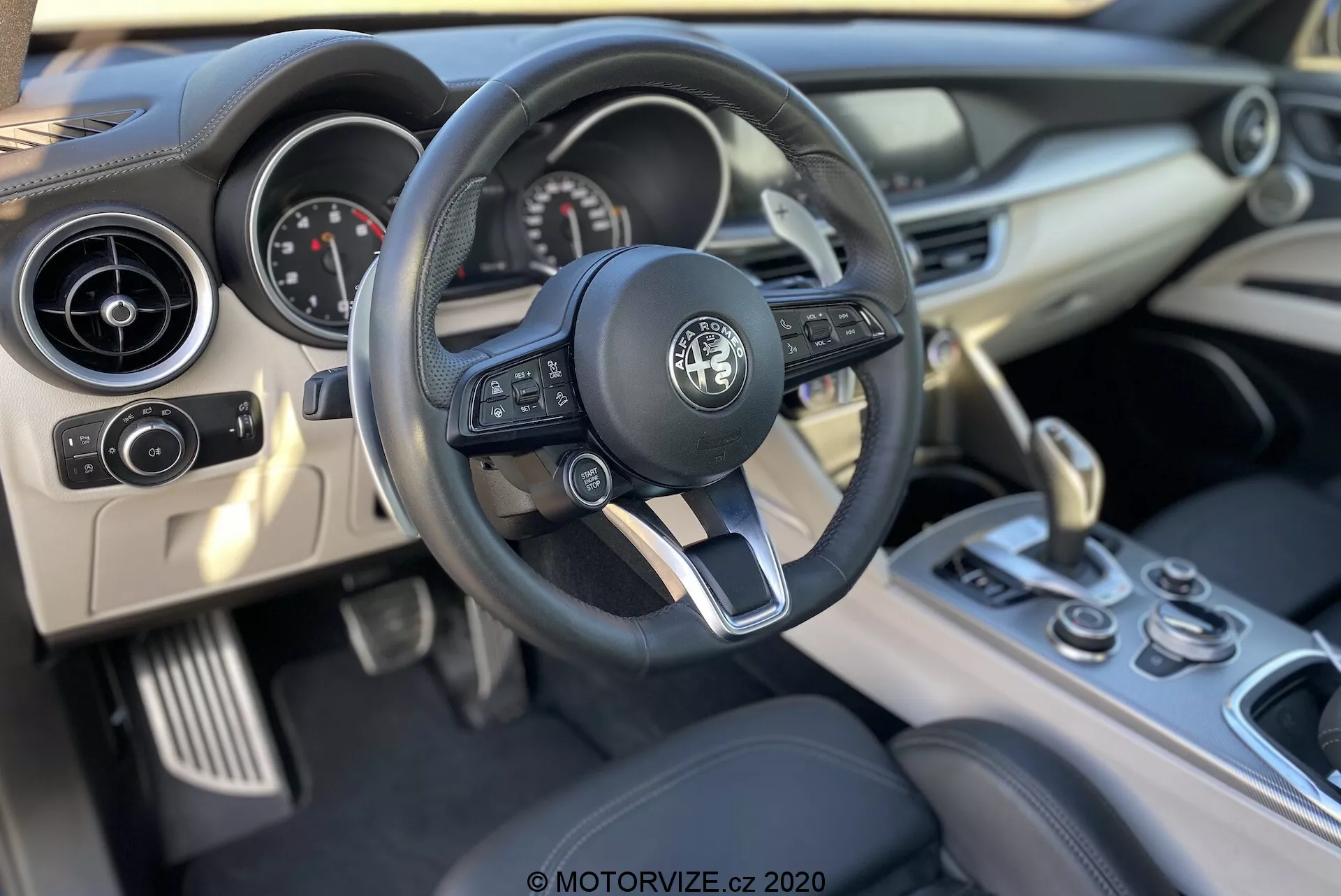 Vista interior del Alfa-Romeo Stelvio 2017 desde la perspectiva del conductor, destacando el volante con la insignia Alfa Romeo, controles integrados de audio y crucero, levas de cambio, tacómetro y velocímetro analógicos, pantalla digital, ventilaciones de aire circulares, salpicadero de tacto suave, botones en la consola central, dial giratorio y selectora de cambio de cuero.