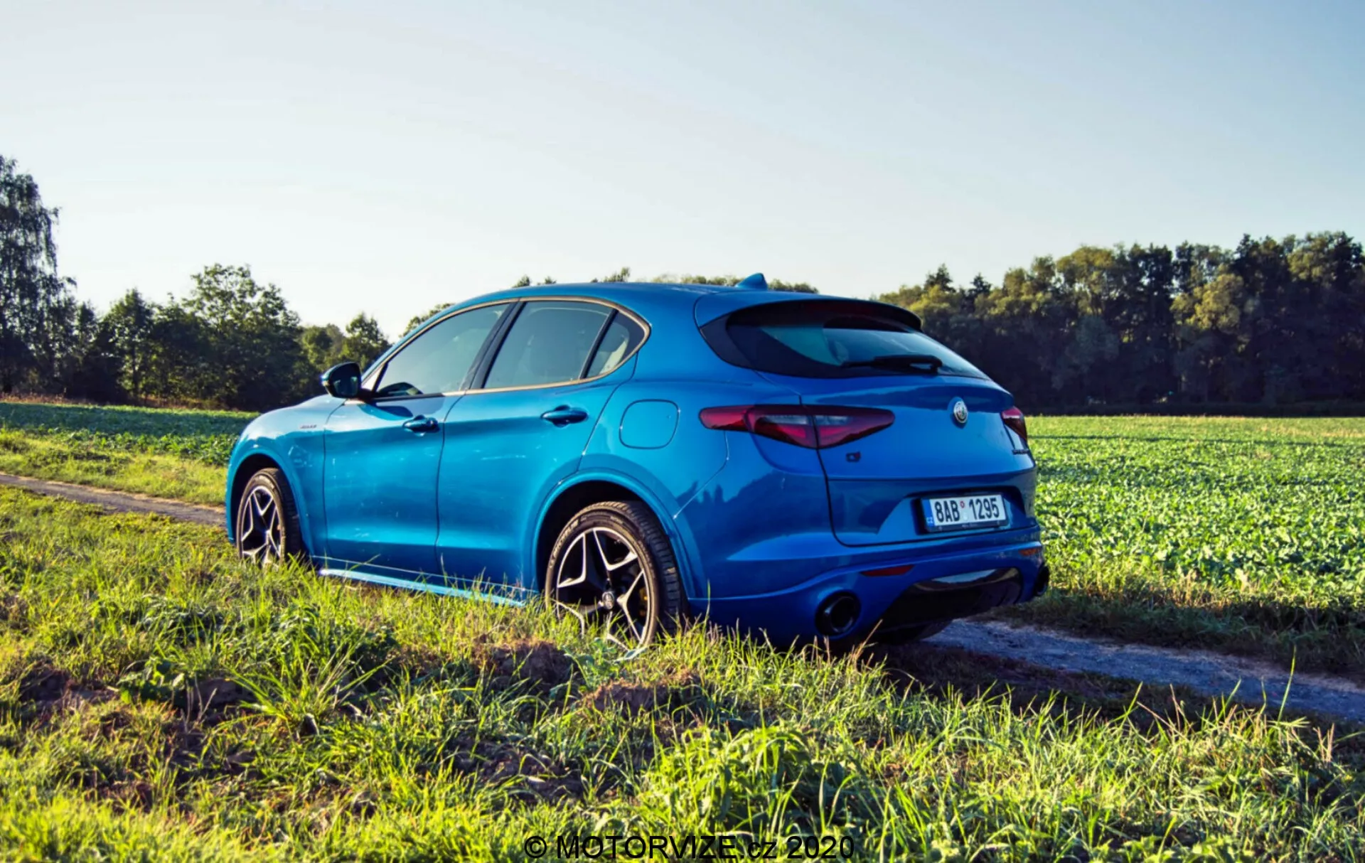 Imagen de un Alfa Romeo Stelvio azul de 2017, un SUV crossover deportivo moderno, tomada desde una perspectiva trasera de tres cuartos en un camino de tierra adyacente a un campo durante la hora dorada. Las características incluyen un alerón deportivo, luces traseras elegantes, difusor trasero con doble salida de escape, placa de registro central y emblema. La vista lateral muestra arcos de rueda pronunciados, línea característica a través de las puertas, grandes ruedas de aleación multirradio con neumáticos de perfil bajo, espejos laterales del color de la carrocería y una antena trasera. El Stelvio tiene cristales tintados en las ventanas traseras, una fuerte línea de hombro y un efecto de destello de luz del sol que resalta su diseño dinámico.