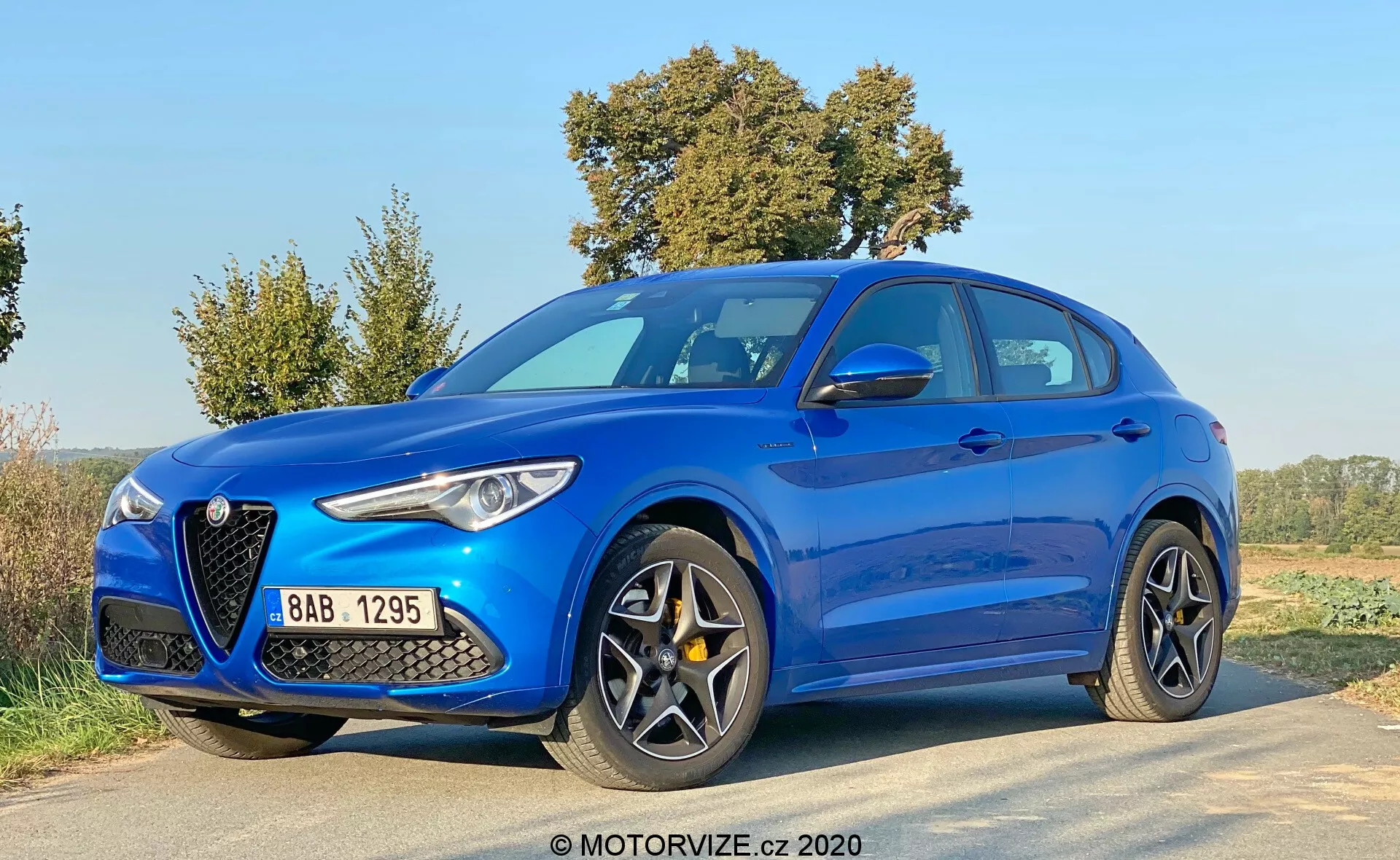 Alfa Romeo Stelvio 2017 azul, fotografiado desde un ángulo de tres cuartos frontal bajo, mostrando su distintiva parrilla en forma de escudo, faros alargados y elegantes, grandes entradas de aire, líneas contorneadas del capó y elegantes llantas de aleación de varios radios en una carretera rural.