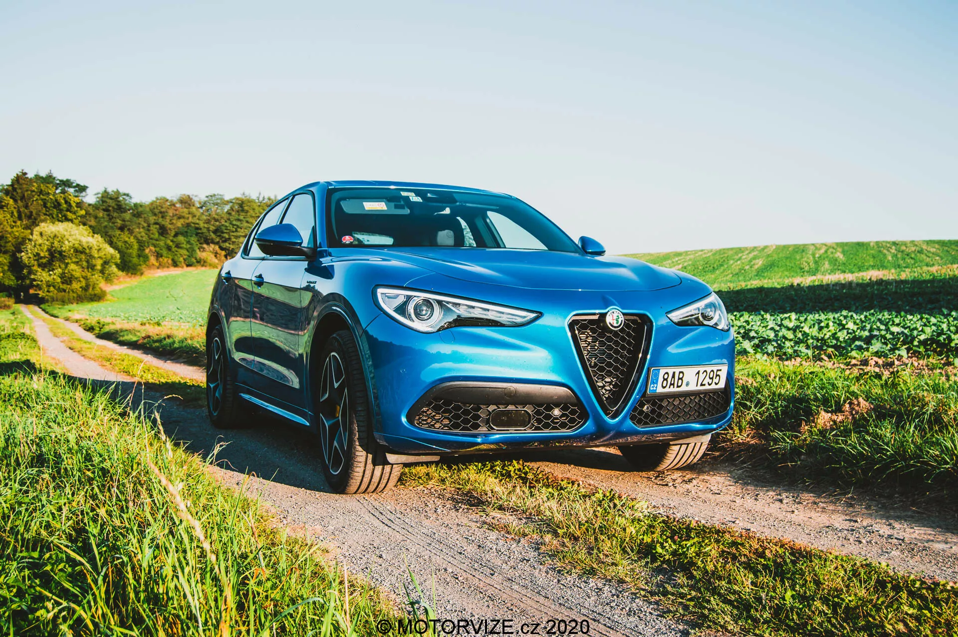 Alfa Romeo Stelvio 2017 en azul estacionado sobre un camino de grava junto a un campo en la tarde, mostrando la parrilla frontal con borde cromado, faros LED, líneas esculpidas del capó y ruedas de aleación multirradiales desde un ángulo frontal de tres cuartos.