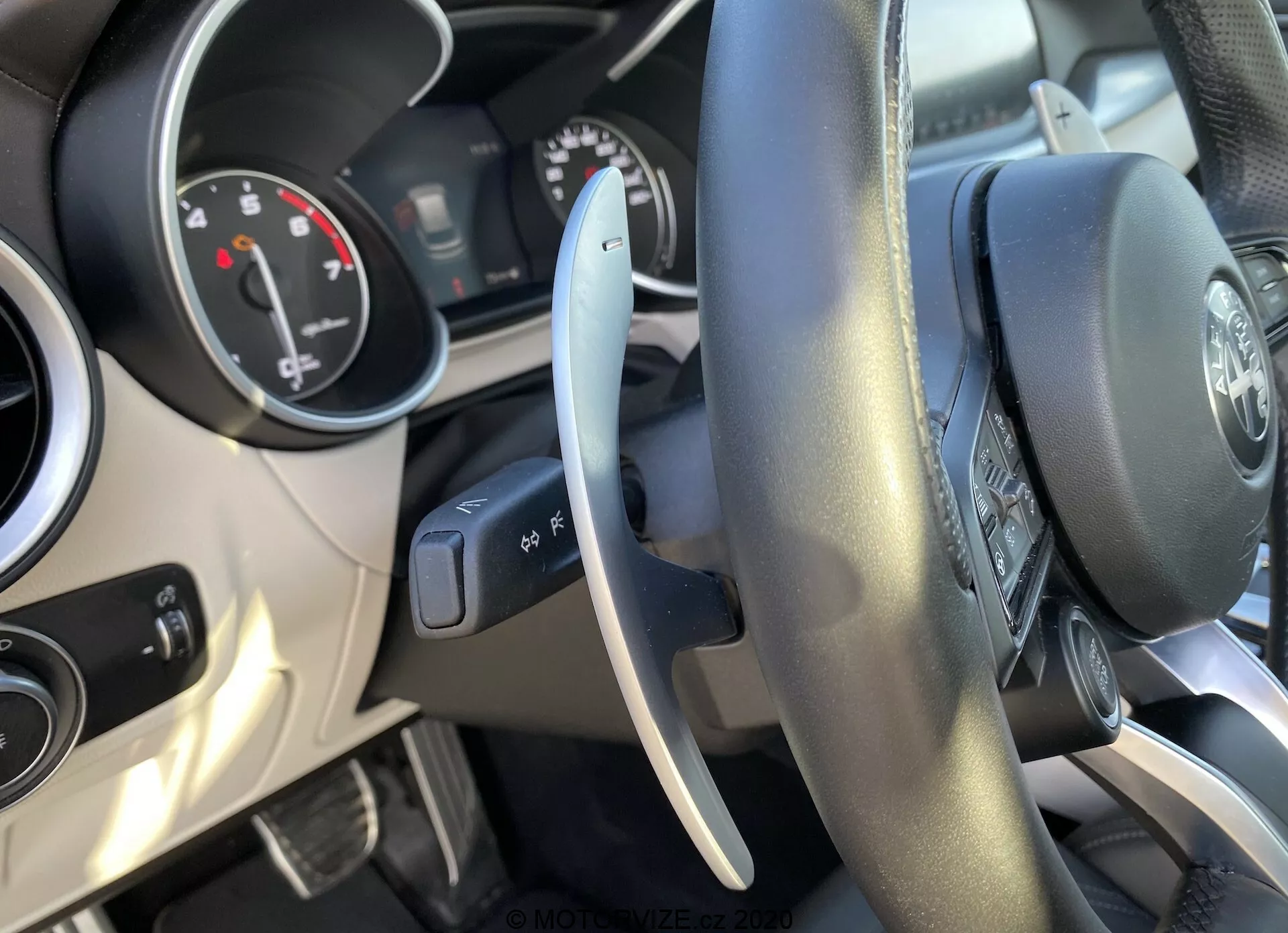 Vista interior de un Alfa-Romeo Stelvio 2017 desde el asiento del conductor, destacando el volante revestido de cuero con botones multifunción, cuadro de instrumentos parcialmente visible con diales analógicos y pantalla digital central, y palanca de control a la izquierda para las luces exteriores. El salpicadero cuenta con ventilaciones centrales con acentos cromados. Imagen bien iluminada que muestra la interfaz y los controles del conductor.