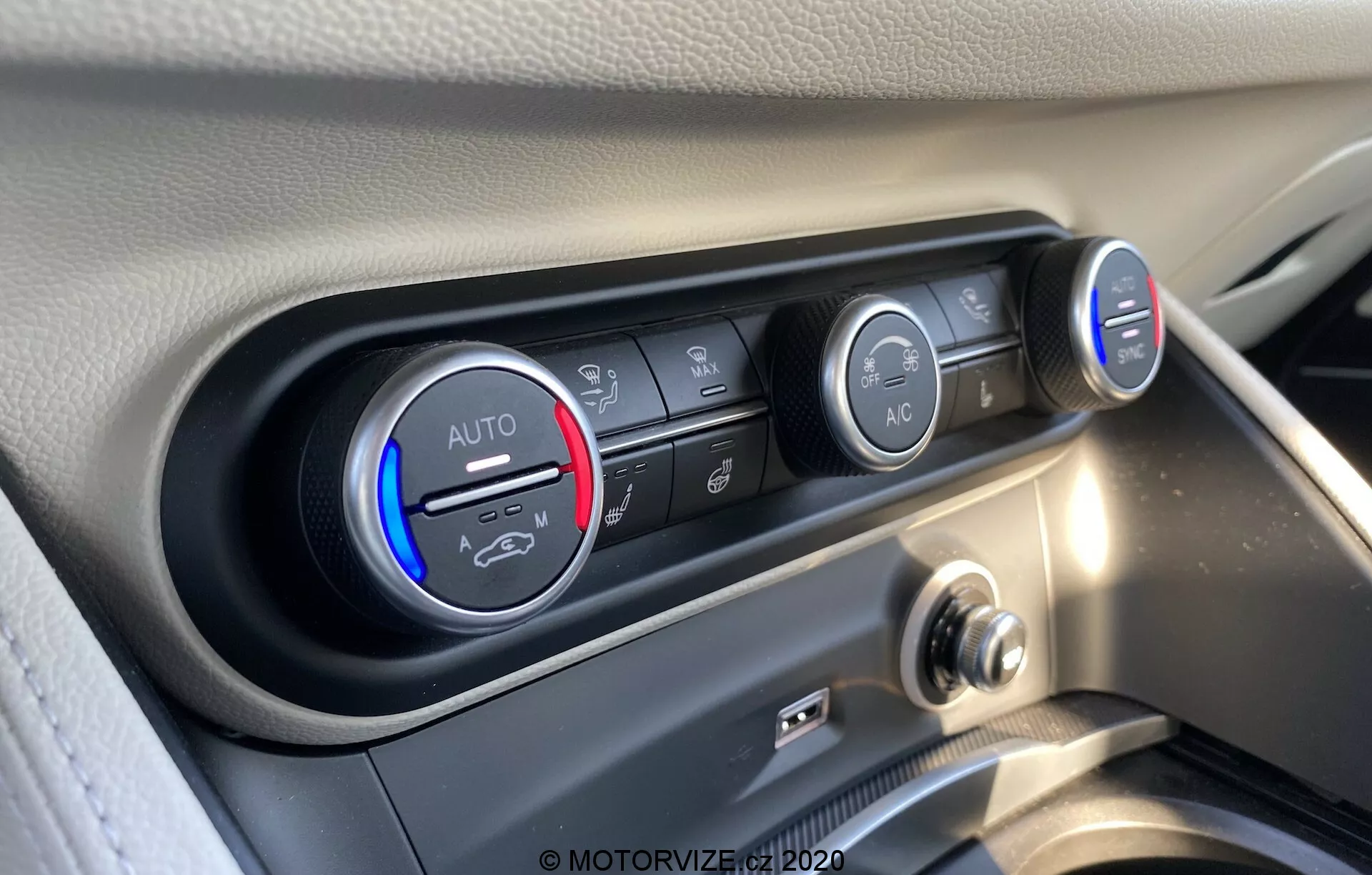 Vista de primer plano del sistema de control de clima del interior del Alfa Romeo Stelvio 2017. La imagen muestra los controles de temperatura y ventilador desde un ángulo ligeramente elevado, enfocándose en los detallados botones y perillas. La perilla de temperatura, ajustada en frío, presenta una banda azul y roja, marcada con 'AUTO'. Al lado, múltiples botones horizontales controlan la dirección del flujo de aire, la recirculación del aire y el desempañado del parabrisas. La perilla derecha indica la velocidad del ventilador con luces LED, ajustada a baja. Debajo, destaca un botón 'A/C'. El panel de control está enmarcado en un entorno negro brillante, contrastando con el tablero claro, capturado en alta resolución con reflejos claros de luz natural.
