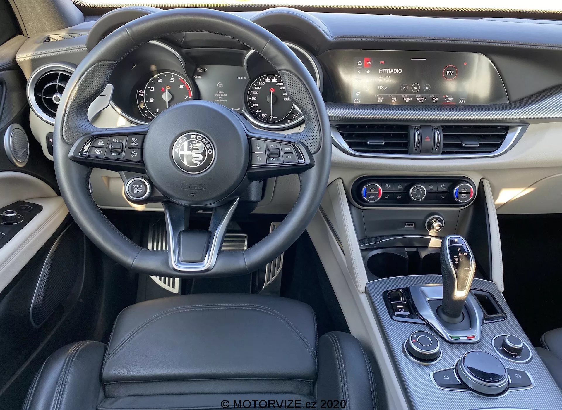 Imagen del interior de un Alfa Romeo Stelvio 2017, centrada en la zona del conductor que muestra el volante multifuncional moderno con forro de cuero y varios botones de control, un cuadro de instrumentos analógico-digital parcialmente visible y la consola central con sistema de infoentretenimiento táctil y control de clima. El diseño combina lujo y deportividad con tapicería de cuero y costuras en contraste. La palanca de cambios y el selector de modos de conducción resaltan la transmisión automática y los modos de conducción seleccionables del vehículo.