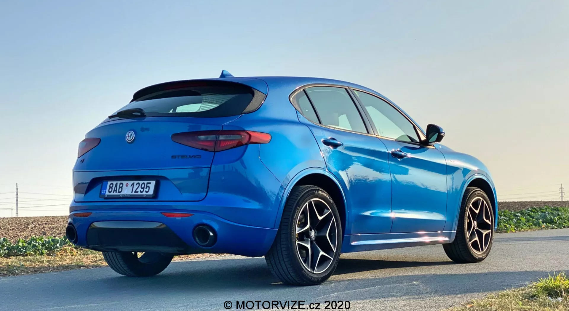 Vista de tres cuartos trasera de un SUV compacto azul Alfa Romeo Stelvio 2017 estacionado al lado de una carretera durante el día con cielo despejado, mostrando alerón trasero deportivo, luces traseras LED, sistema de escape doble, grandes ruedas de aleación con múltiples radios, sensores de estacionamiento y antena de aleta de tiburón, en un entorno que resalta su diseño elegante y características modernas.