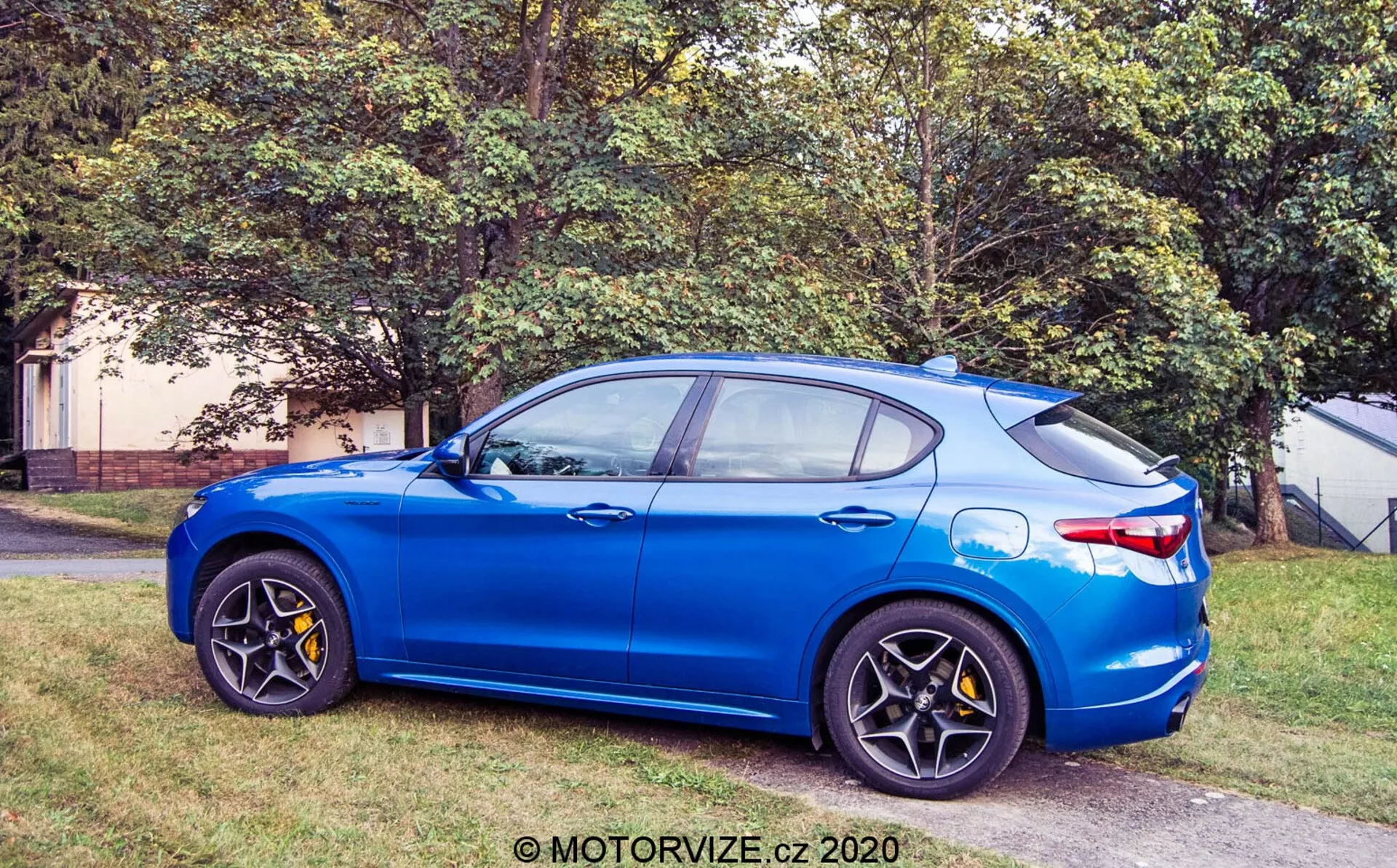 Vista de perfil lateral del Alfa Romeo Stelvio 2017 en azul brillante aparcado en un área verde junto a una superficie pavimentada, mostrando la vista del cuarto delantero con llantas de aleación negras de múltiples radios, pinzas de freno amarillas, espejos laterales del color de la carrocería, línea del techo inclinada con antena de aleta de tiburón, y diseño de cuatro puertas con acabado metálico.