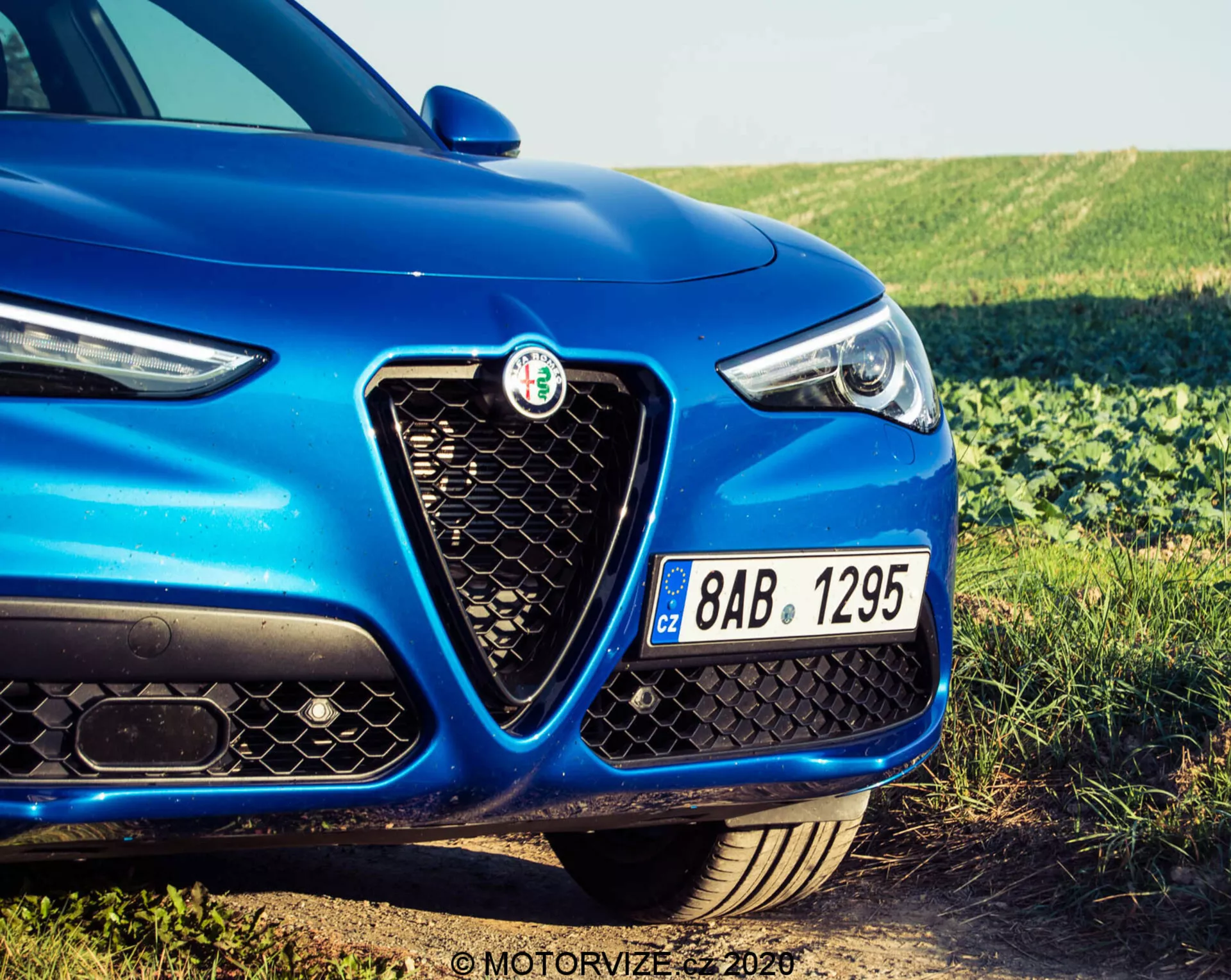 Primer plano del lado izquierdo delantero de un Alfa Romeo Stelvio 2017 azul, mostrando el distintivo emblema en la parrilla hexagonal, el diseño elegante del faro con elementos LED y parte del capó. La imagen, tomada desde un ángulo bajo, resalta la fascia delantera deportiva y agresiva del vehículo, que incluye el parachoques delantero negro con grandes tomas de aire y una matrícula checa.