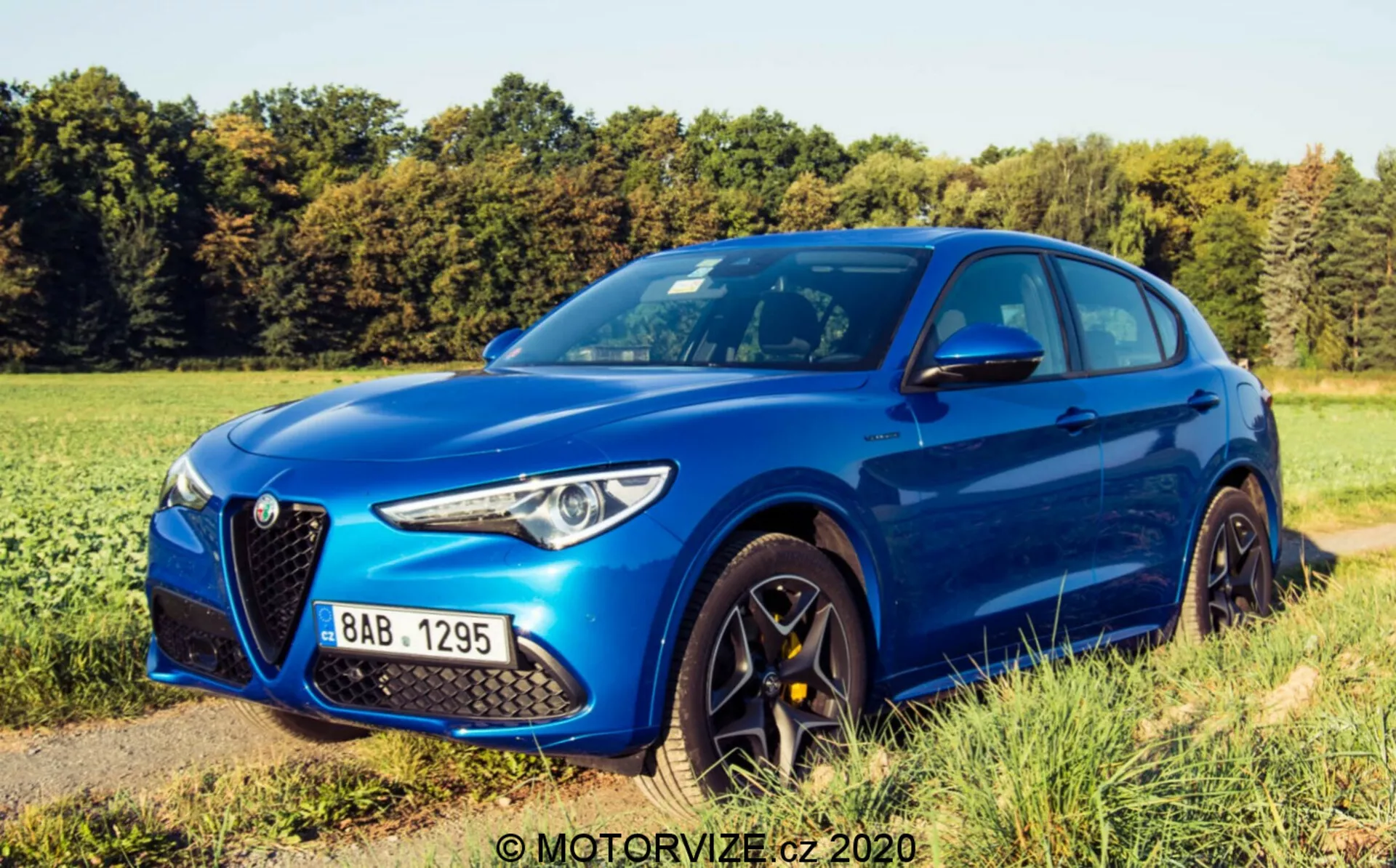 Vista de un ángulo de tres cuartos delantero del Alfa Romeo Stelvio 2017 en azul metálico vibrante, destacando la parrilla triangular 'Scudetto' característica, los elegantes faros LED, el diseño aerodinámico del capó con líneas definidas, las llantas de aleación de varios radios de color oscuro, los neumáticos de alto rendimiento de perfil bajo, los espejos laterales del color de la carrocería con señales de giro integradas y los marcos de las ventanas del color de la carrocería con el pilar B negro. El vehículo está aparcado en un camino sin pavimentar al lado de un campo de hierba bajo un cielo despejado con sombras suaves, lo que sugiere luz de temprano en la mañana o tarde en la tarde.
