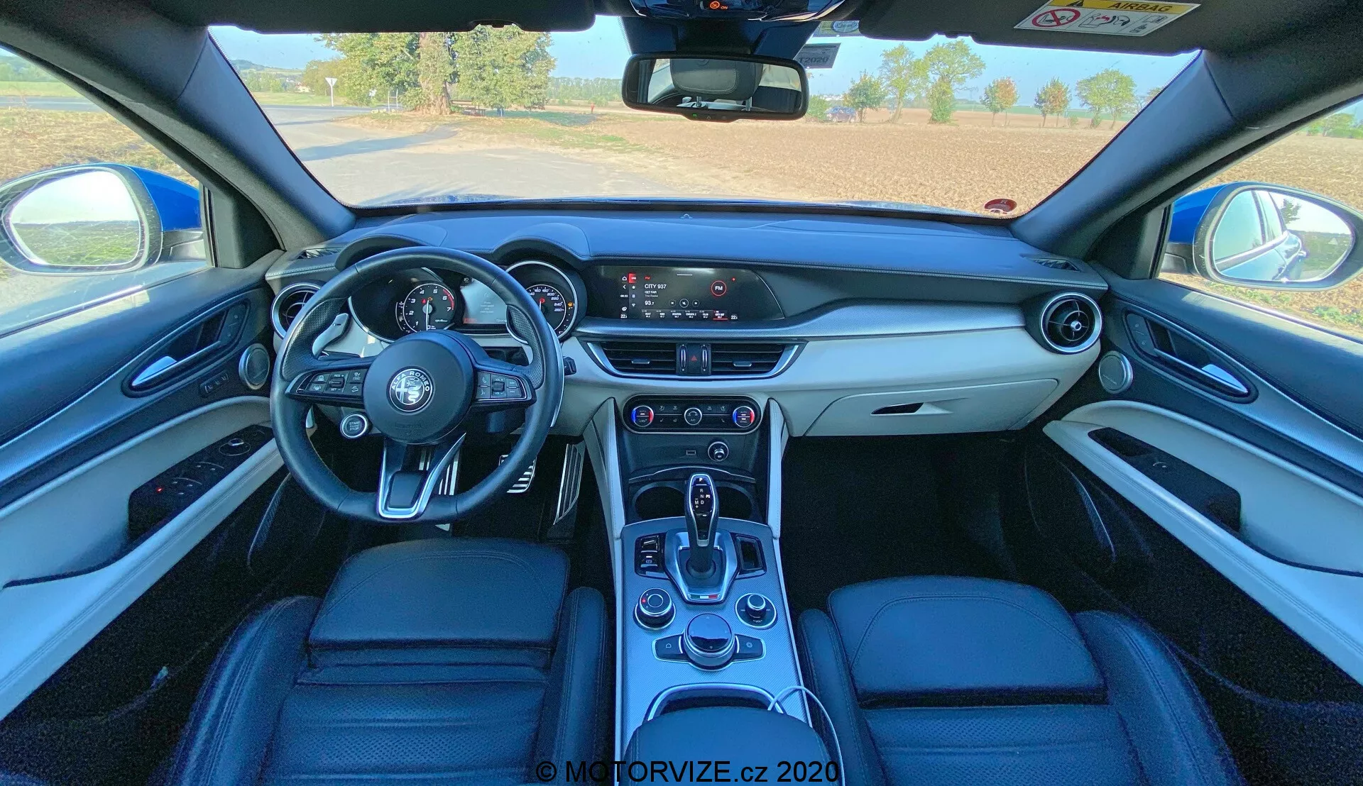 Vista interior de un Alfa Romeo Stelvio 2017 desde una perspectiva en primera persona, que muestra el tablero, volante de tres radios con controles montados, centro de información digital del conductor, sistema de infoentretenimiento con pantalla táctil, consola central con palanca de cambios automática y tapicería de cuero premium con materiales contrastantes.