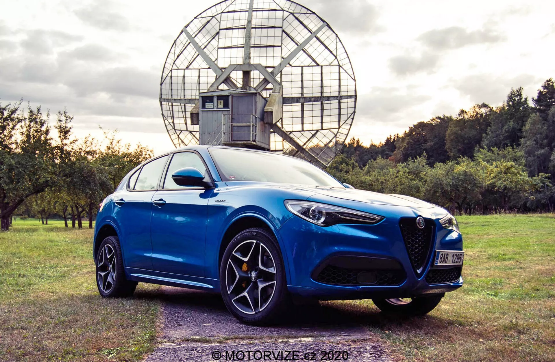 Foto de un sedán deportivo moderno azul capturada desde un ángulo bajo de tres cuartos delanteros, mostrando el perfil frontal y lateral. El coche cuenta con una parrilla triangular distintiva, faros afilados y angulares, y llantas de aleación multiradio con pinzas de freno visibles. Está aparcado en una zona de césped con árboles y una estructura esférica de aspecto industrial en el fondo. La toma está compuesta bajo luz natural, resaltando las líneas aerodinámicas y el diseño suave del coche.