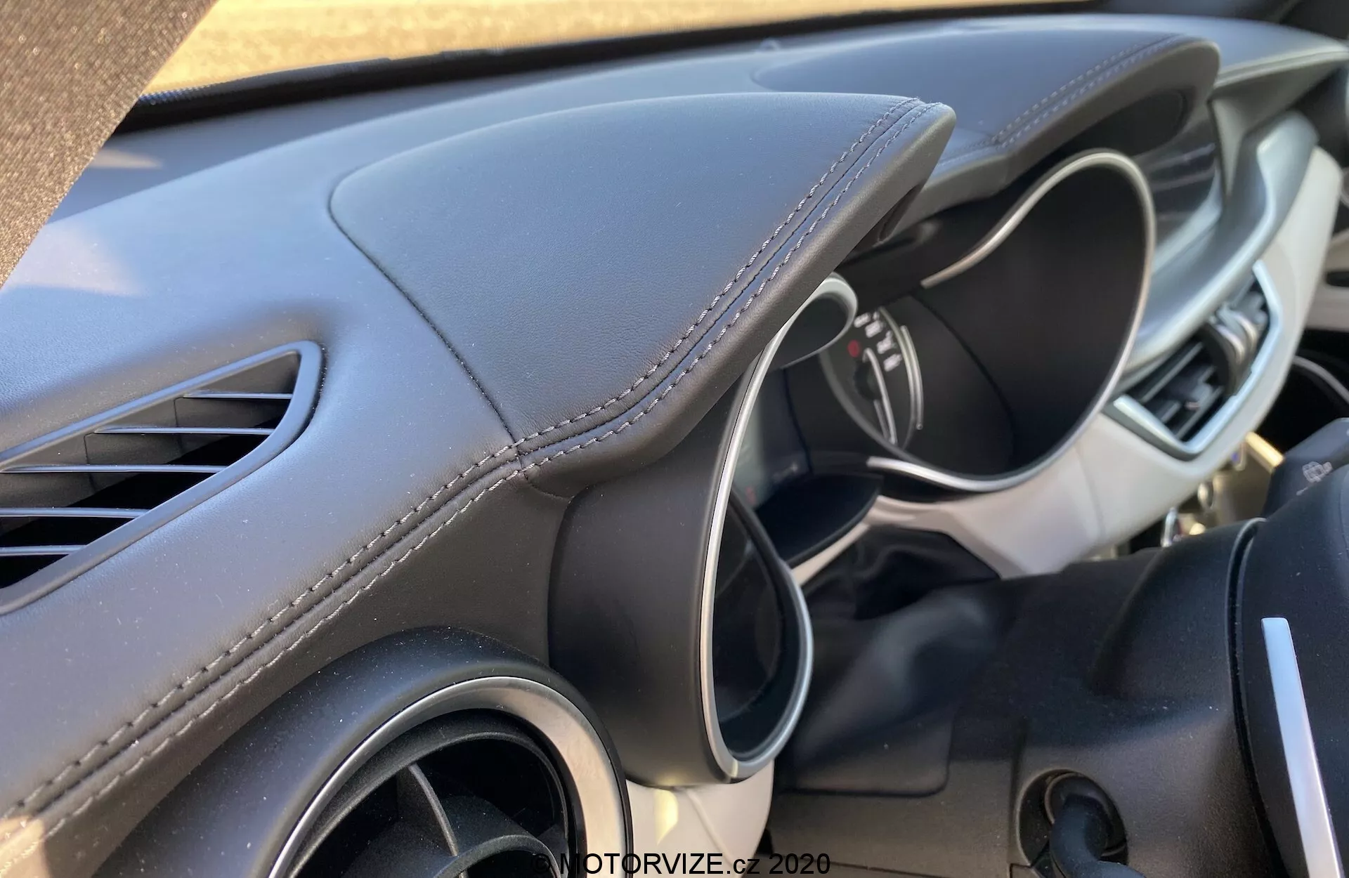 Vista de cerca del interior del Alfa-Romeo Stelvio 2017, enfocándose en el lado del conductor del tablero, visible desde el asiento del pasajero o la consola central. El diseño moderno y esculpido del tablero presenta material suave al tacto con costuras blancas, sugeriendo una estética premium. También se puede ver la rejilla de ventilación izquierda y parte del volante multifuncional con controles o botones, lo que sugiere un entorno avanzado y fácil de usar para el conductor.