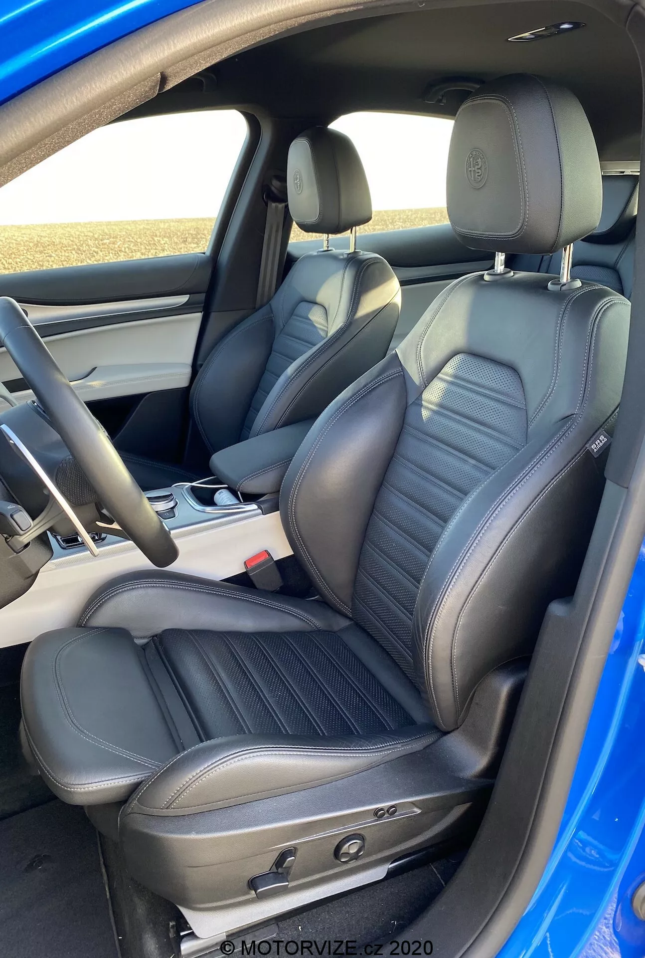 Interior del Alfa-Romeo Stelvio 2017 destacando el área moderna de la cabina delantera con asientos delanteros tapizados en cuero negro con diseño de panel central ribeteado, reposacabezas integrados, soporte lumbar ajustable y controles electrónicos para asientos ajustables eléctricamente. La imagen, tomada desde la perspectiva de alguien parado justo fuera del lado del pasajero con un ángulo ligeramente hacia abajo, muestra la consola central, compartimento de almacenamiento detrás de la palanca de cambios, volante con controles montados, paneles de puertas negros a juego con manijas plateadas cepilladas y botones de control de ventanas eléctricas, cinturones de seguridad de tres puntos retráctiles estándar y un pilar B que indica la separación de las cabinas delanteras y traseras.