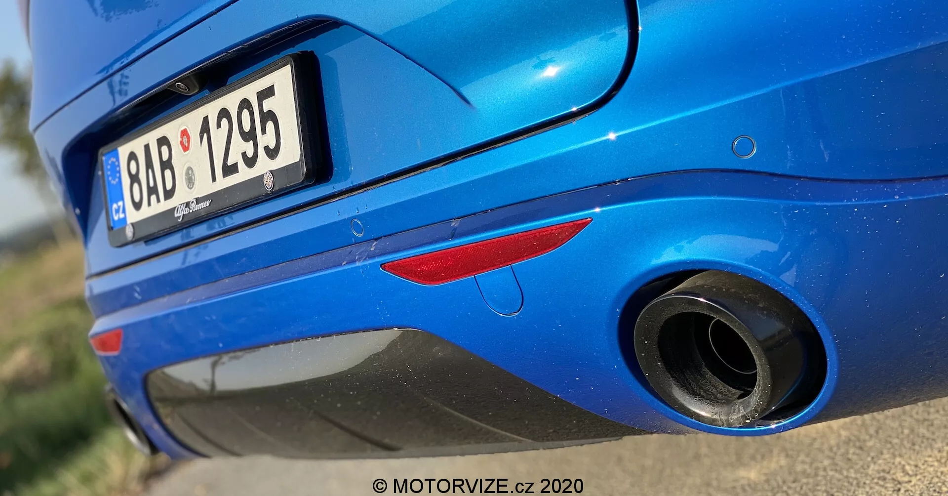 Vista en primer plano de la sección trasera de un Alfa Romeo Stelvio 2017 azul, tomada desde un ángulo bajo. La toma resalta la mitad inferior de la parte trasera del coche, centrándose en el tubo de escape y el parachoques trasero. Las características incluyen una luz trasera reflectora roja, un neumático trasero parcialmente visible, un parachoques trasero redondeado y brillante, una placa de matrícula parcialmente recortada, un tubo de escape redondeado con punta de cromo y un pliegue sobre el escape en el parachoques. La pintura metálica con reflejo de la luz del sol enfatiza la condición limpia y bien mantenida del coche.