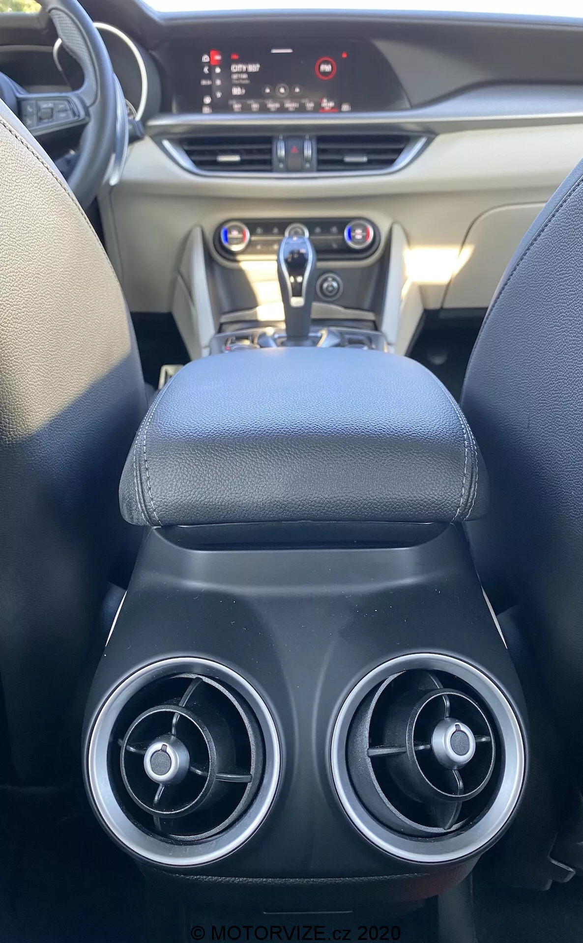 Vista interior desde el asiento trasero de un Alfa Romeo Stelvio 2017, enfocándose en la consola central y el tablero. La imagen muestra dos salidas de aire circulares, un reposabrazos de cuero, una palanca de cambios automática, panel de control brillante con controles metálicos y un tablero digital iluminado, destacando el diseño elegante y funciones modernas.
