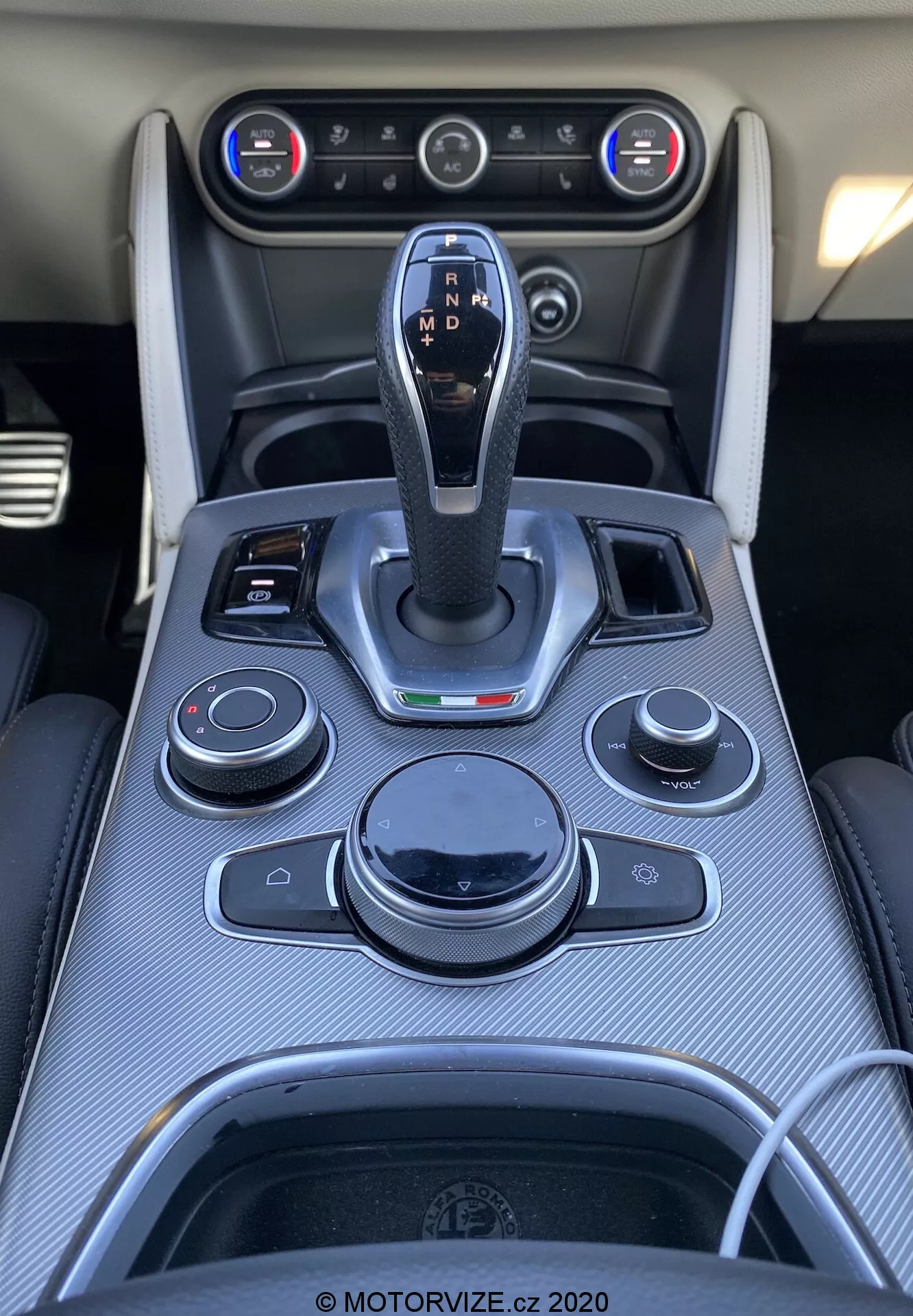 Vista de primer plano hacia abajo del centro de la consola en un interior de Alfa Romeo Stelvio 2017. Palanca de cambios central con acabado de cuero y detalles en cromo, etiquetada con configuraciones de transmisión automática. Mandos y botones circundantes para el sistema de infoentretenimiento, audio, clima y funciones del vehículo. Moldura de fibra de carbono, ventilas de aire acondicionado y sistema de control climático de doble zona visibles. Porta vasos con contorno de cromo y un puerto de conectividad presente.