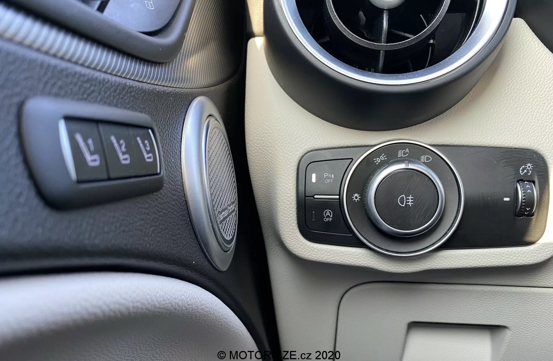 Primer plano del módulo de control de luces y el tablero del Alfa Romeo Stelvio 2017 desde la perspectiva del conductor o pasajero delantero. La imagen muestra los controles basados en perillas para los faros, luces de estacionamiento, iluminación automática y luces antiniebla delanteras y traseras. Adyacente se encuentra una palanca para ajustar el ángulo de los faros. Los elementos visibles incluyen una ventilación circular, un tablero bicolor en negro y beige, botones para el control de tracción, liberación del maletero, asistencia de estacionamiento, un emblema circular probablemente representando la marca del coche o el sistema de audio y una rejilla de altavoz redondeada.